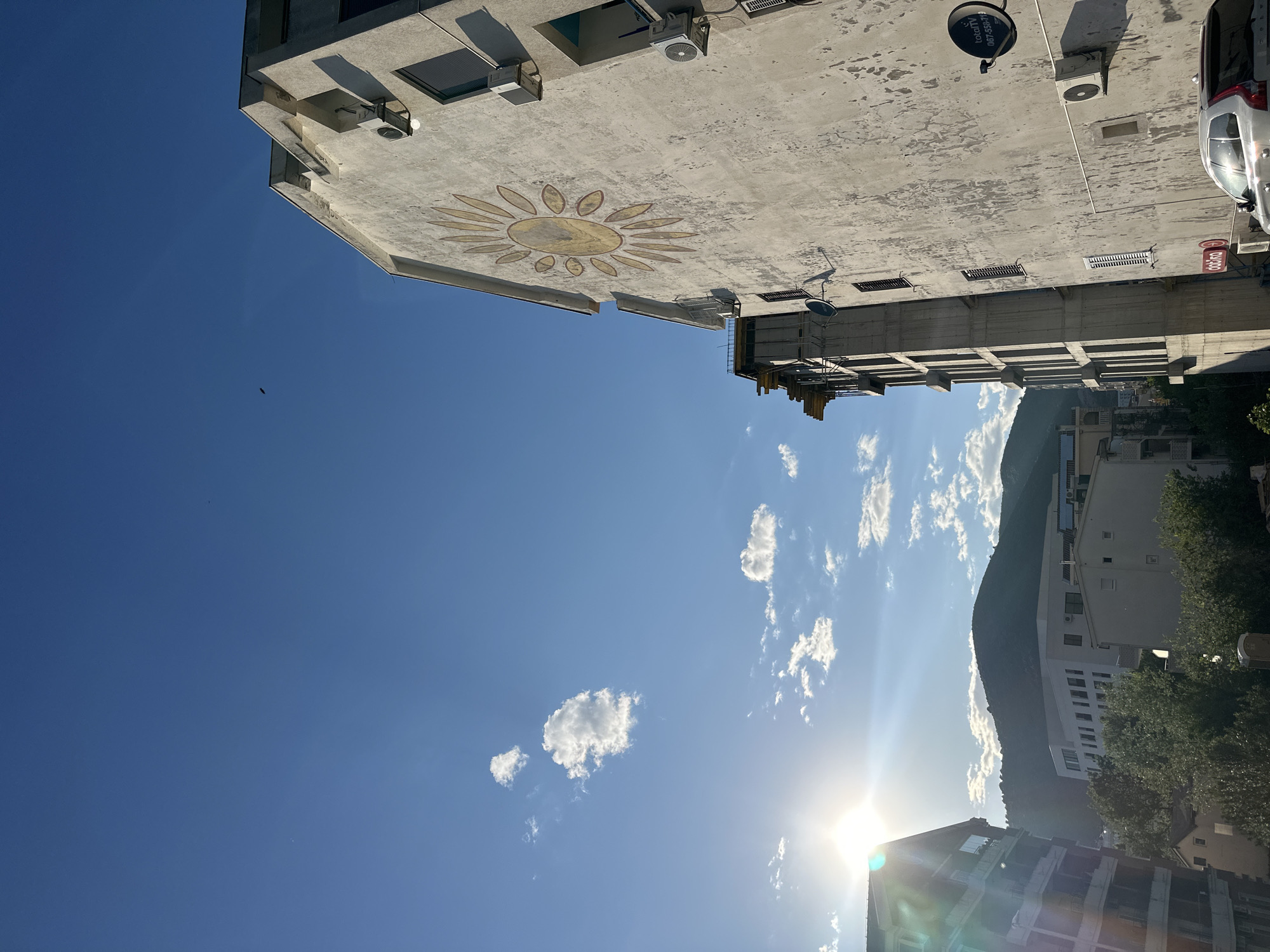 The image presents a bright urban landscape under a clear blue sky, likely in Budva, Montenegro. The view looks upwards, with the sun shining intensely from the lower right, creating a prominent lens flare and illuminating scattered white cumulus clouds across the sky. A tiny dark speck, possibly a bird, is visible in the upper-left sky.
On the left side, a large, light-colored concrete building dominates the foreground, its facade textured and showing signs of age. A distinctive, circular yellow sun design with radiating petals is painted on one of its prominent walls. Several air conditioning units and rectangular vents are attached to the building's exterior.
To the right, other buildings, including what appears to be part of a residential or commercial block, extend into the mid-ground. A white car is partially visible parked on the far right. A black satellite dish, affixed to one of the buildings, displays the text "totalTV", a phone number "087-353018", and "www.totaltv.tv". Below this, a small red sign features the Thai script "รอยยิ้ม" (roy yim), which translates to "smile".
In the background, a large, lush green mountain range rises against the horizon, characteristic of coastal Mediterranean regions. The bright, high sun suggests the photo was taken during late morning or early afternoon. The scene is static, devoid of people, and conveys a sense of a warm, sunny day in a vibrant town.