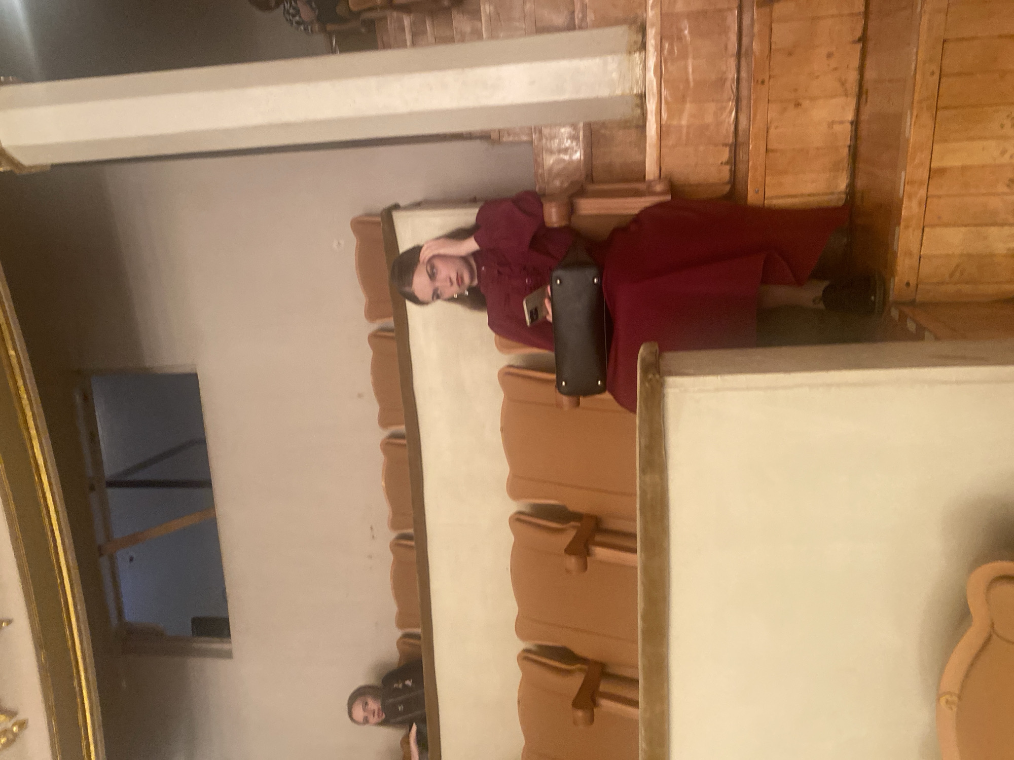 The image shows a woman in a red dress sitting in an audience seating area that may be in a theatre or cinema. She is holding a mobile phone and a handbag, and a second person is partially visible in the lower left. The location is potentially in the city of Yekaterinburg, Russia. The seating consists of wooden benches. There is wooden paneling on the wall. The lighting appears low, creating a slightly dim atmosphere.