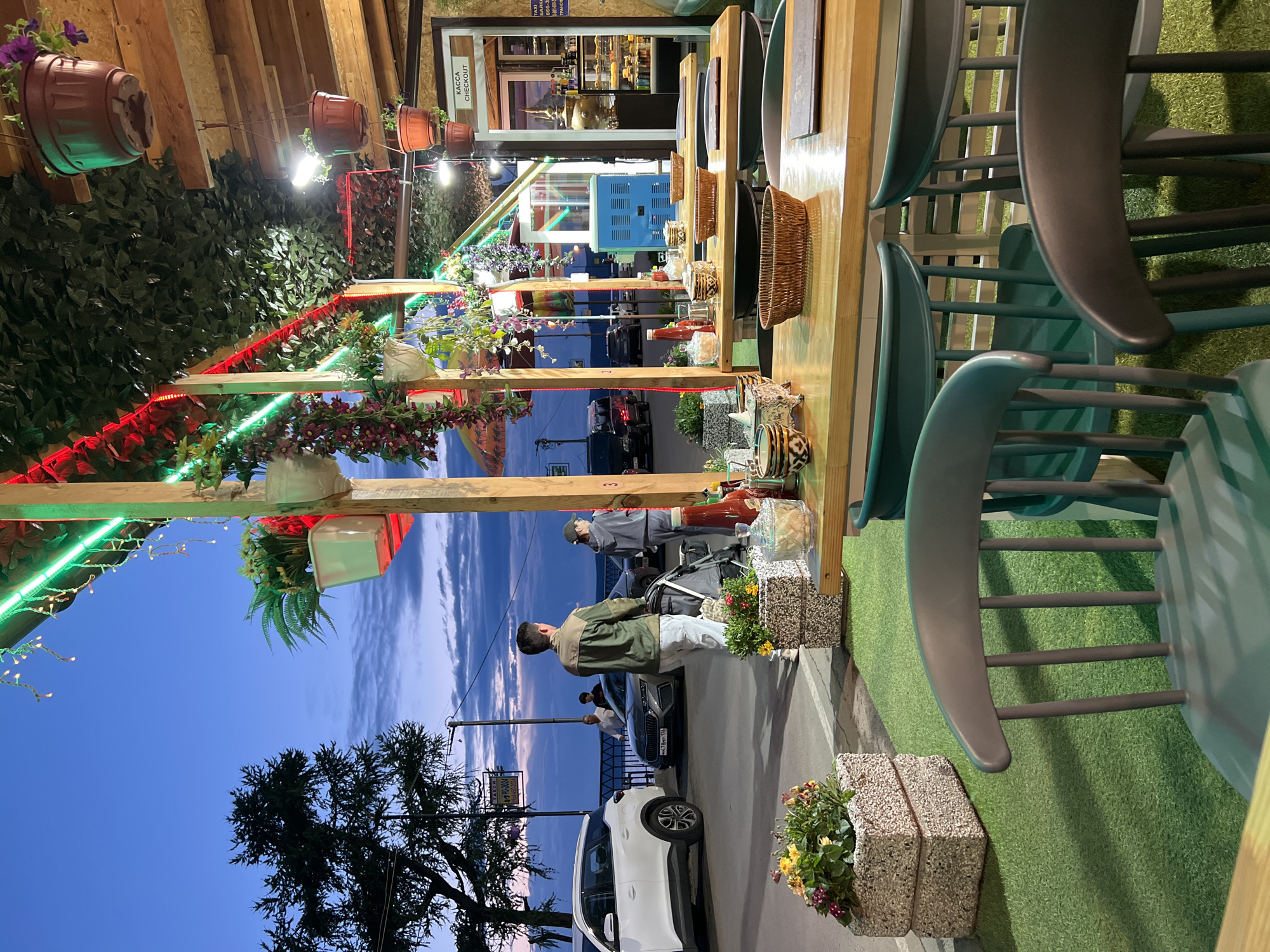 An outdoor seating area of a cafe in Listvyanka, Russia, is depicted at dusk. The foreground features green artificial turf and several empty chairs. Beyond the seating area, wooden tables are set with woven baskets and condiments. Further back, a checkout counter is visible through a doorway, displaying goods and a traditional samovar. Outside the cafe, a street runs alongside a body of water, possibly Lake Baikal, with cars and a few pedestrians present. The sky is a deep blue with some clouds, and decorative lights are strung overhead.