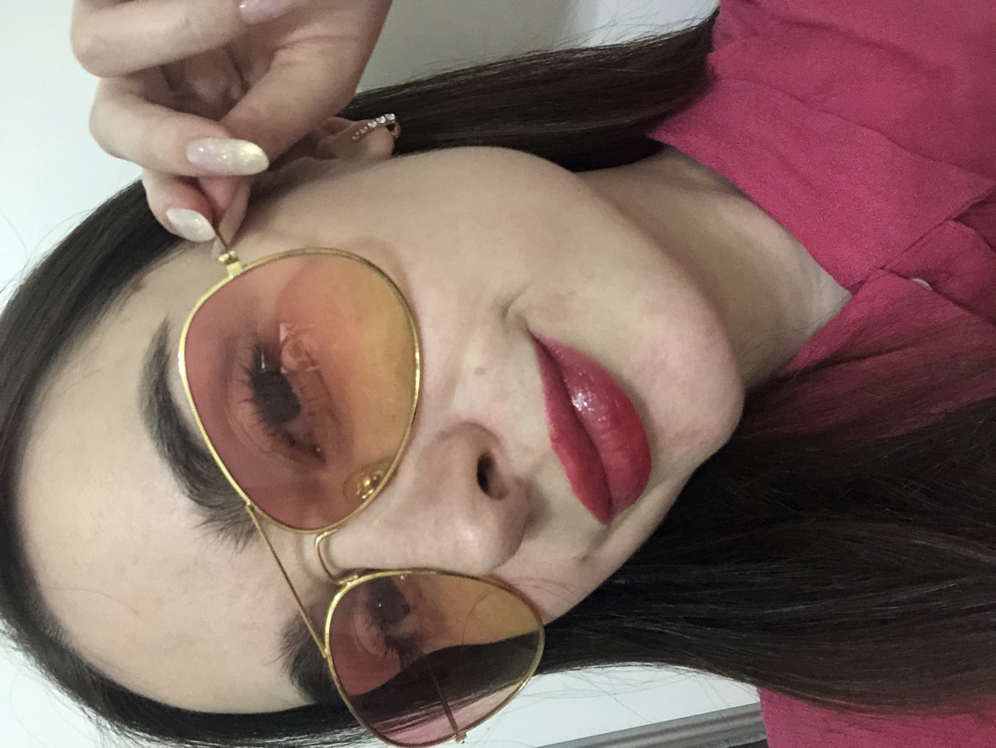 A woman is shown in a close-up, tilted selfie. She is wearing pink tinted aviator sunglasses and red lipstick. Her nails are manicured. She is wearing a red top. There is a ring on her ear. The location is not apparent.