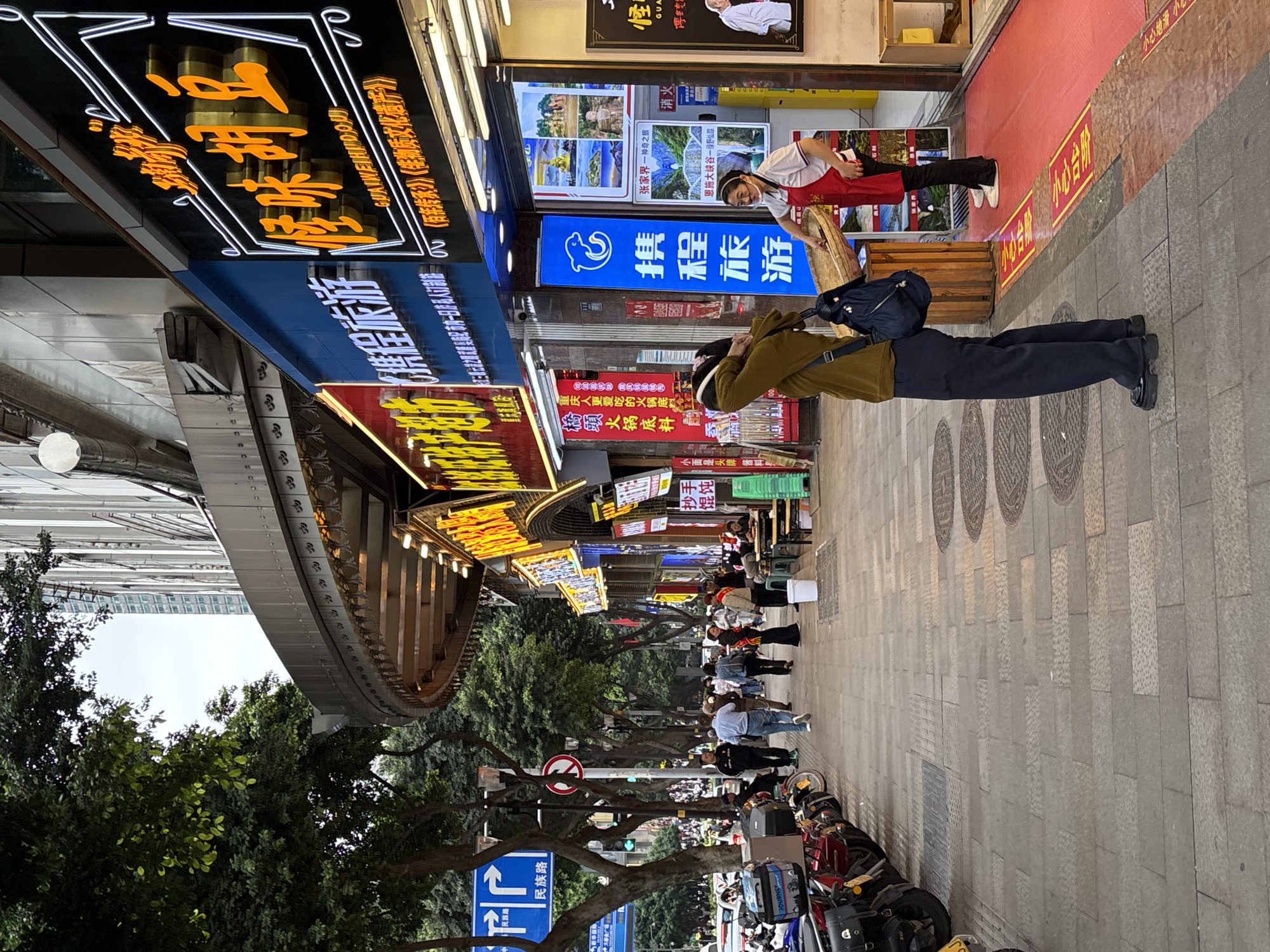 The image shows a street scene in Yuzhong District, China. The scene is a commercial area, with shop signs in Chinese. People are walking along the sidewalk, and there are motorbikes parked along the curb. Signs for businesses and destinations, including Zhangjiajie National Forest Park and Enshi Grand Canyon, are also visible.