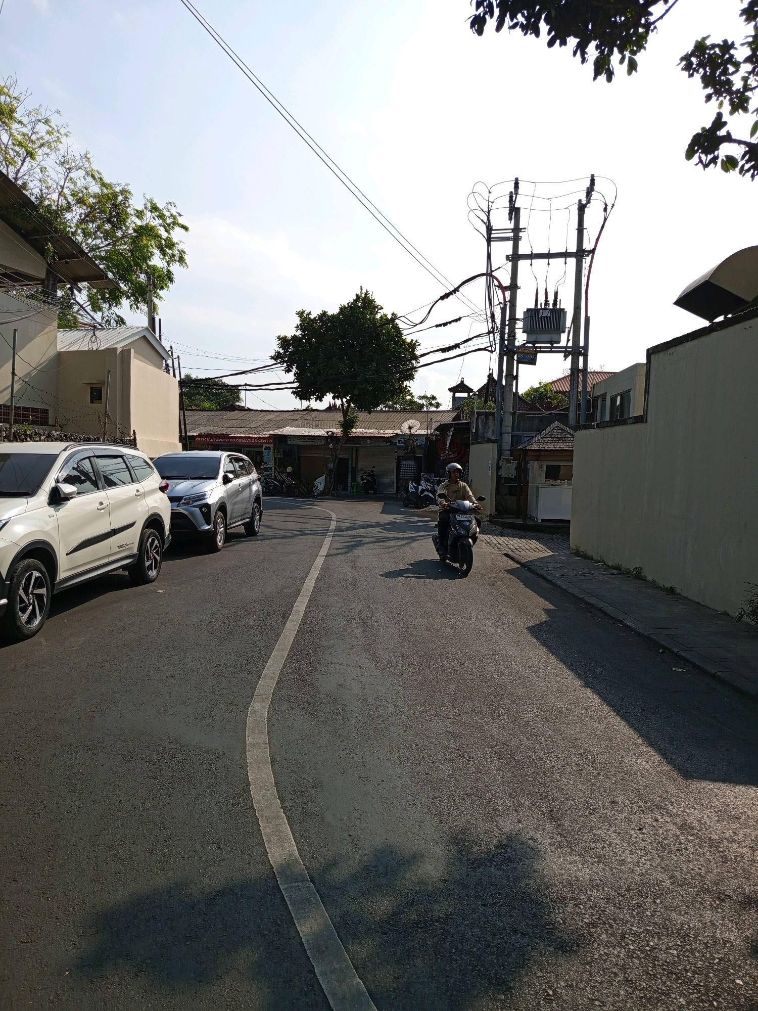 This is a street-level, eye-level, medium shot of a paved road in Denpasar, Indonesia, with buildings and electrical infrastructure lining the sides. The road has a white dashed line marking its edge. Two SUVs are parked on the left side of the road. A person is riding a motorcycle in the right lane, heading away from the camera. Buildings are visible on both sides of the street, with some displaying signage. An electrical transformer is mounted on poles on the right side of the street, with numerous wires crisscrossing overhead. Trees and foliage are scattered throughout the scene. The sky is bright and clear, suggesting daytime. The overall scene depicts a typical urban street environment in Denpasar.