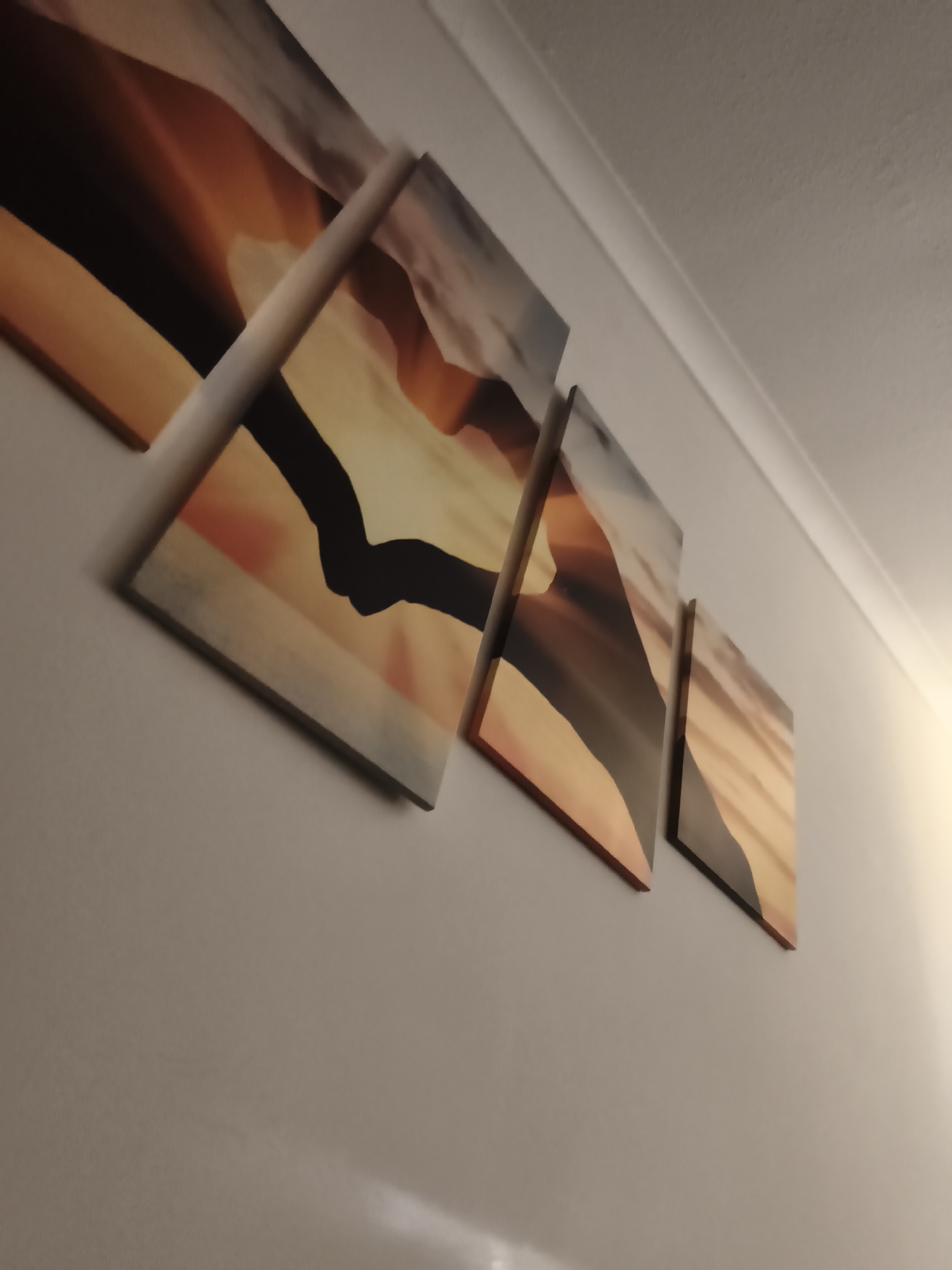 The image captures a close-up, angled view of four multi-panel canvas prints mounted on a light-colored wall within an indoor setting in Huntingdonshire, United Kingdom. The prints are arranged in a staggered, ascending pattern, forming a larger, cohesive artwork. The artwork itself depicts an abstract or stylized landscape, characterized by warm tones of orange, yellow, and brown, with prominent dark, sweeping shapes that suggest dunes, mountains, or contrasting shadows.
The wall is a neutral, light shade, possibly off-white or light grey, and appears smooth. Above the wall, a white coving or cornice is visible, marking the transition to a textured, light-colored ceiling. A warm, soft light illuminates the upper right portion of the scene, casting a glow on the ceiling and wall, suggesting either artificial lighting or natural light filtering in, possibly during evening hours. No people or specific activities are visible, and there is no discernible text. The image focuses on interior decor and the aesthetic display of art.