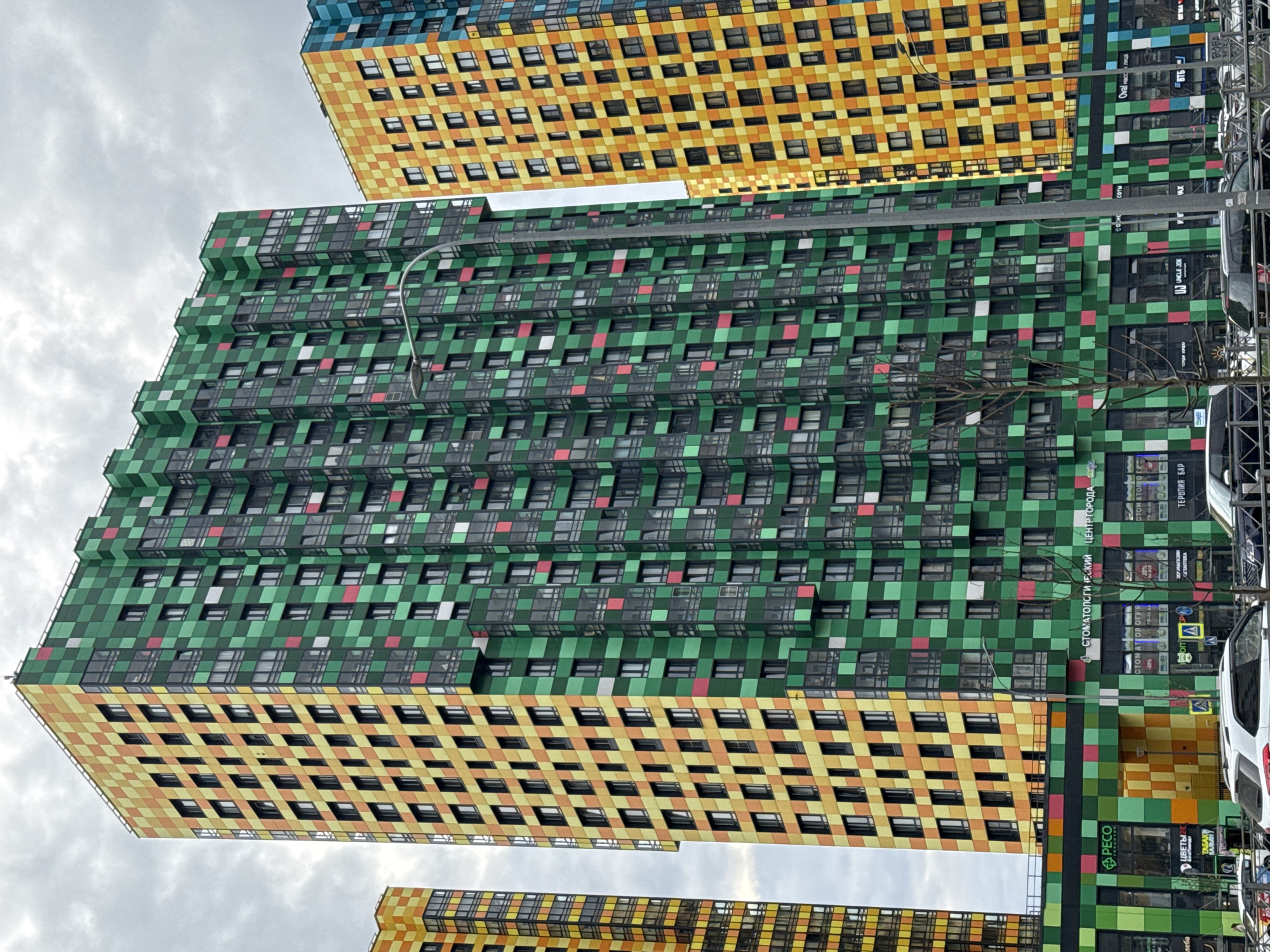 The image shows a close-up, tilted view of modern, colorful apartment buildings in Saint Petersburg, Russia. The buildings feature a blocky, checkerboard pattern with colors like green, yellow, orange, and blue. The buildings have rows of windows. A streetlamp is visible in front of one building. Signage in Russian is visible on the side of one of the buildings, indicating businesses such as a dental center and stores. Cars are parked in front of the buildings, with a white van prominently visible. The sky is cloudy.