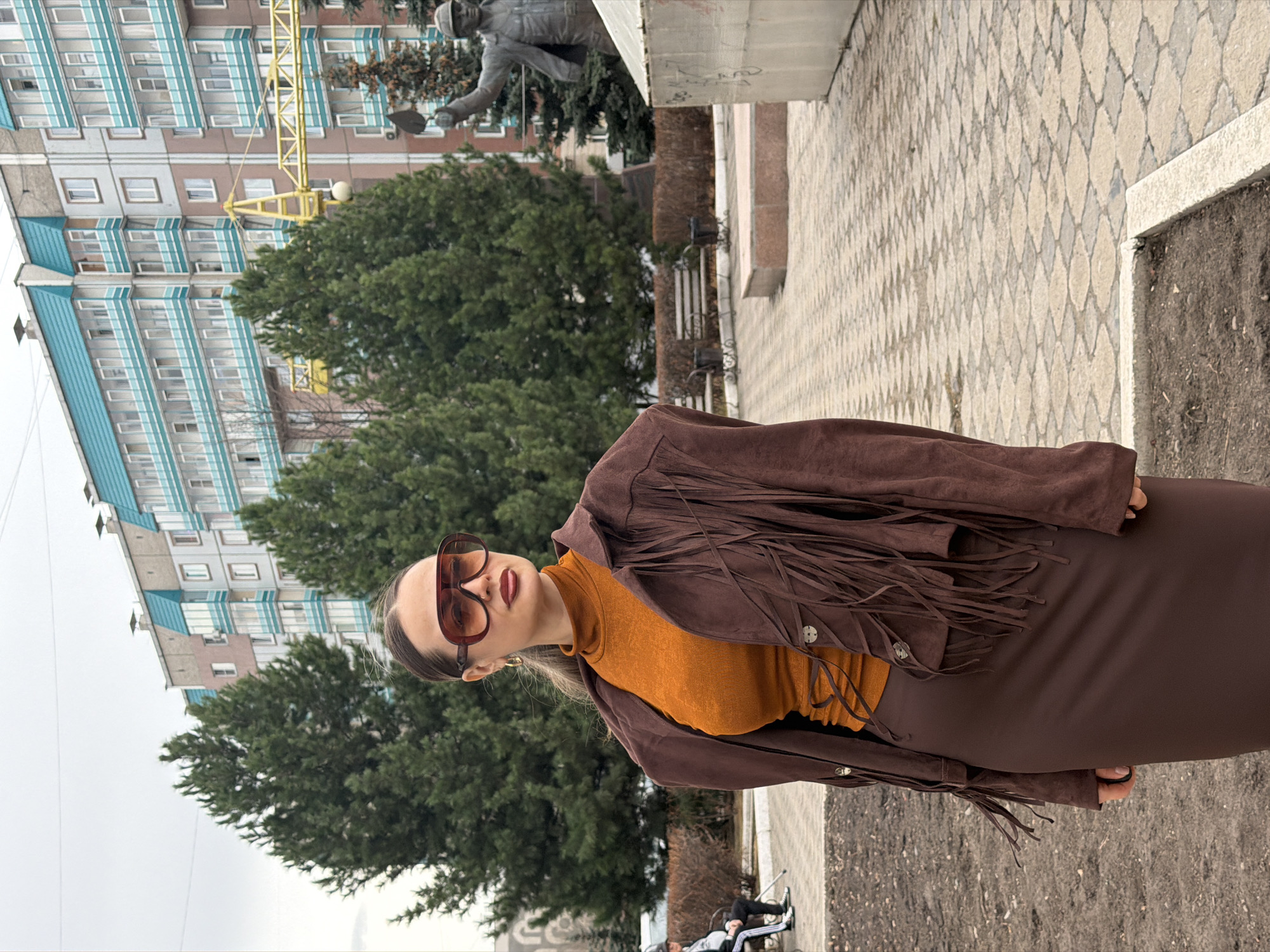 An outdoor portrait shot features a young woman posing in an urban setting. She stands centrally, slightly angled, looking towards the viewer with a neutral expression. She wears large, brown-tinted sunglasses, a snug-fitting orange turtleneck, and a brown suede-like jacket adorned with long fringes across the chest and arms. A dark brown skirt or dress completes her outfit.
The background is rich with urban details. To her left, several large, green deciduous trees are prominent. Behind the trees and extending to the upper left of the frame is a multi-story building, characterized by a repeating pattern of light blue and white cladding on its upper floors and numerous windows. A yellow construction crane is visible in front of this building, partially obscured by the trees. To the right of the woman, a grey statue of a man holding a tool, possibly a trowel or shovel, stands on a pedestal. The ground beneath is paved with light-colored tiles forming a diamond pattern, transitioning to a strip of dark earth on the right. In the bottom left, a bench with two individuals sitting on it is partially visible.
The sky is overcast and uniformly grey, indicating a cloudy daytime. The architectural style of the building and the public statue suggest a post-Soviet urban environment, consistent with the given location context of Krasnoyarsk, Russia. No visible text is present in the image.