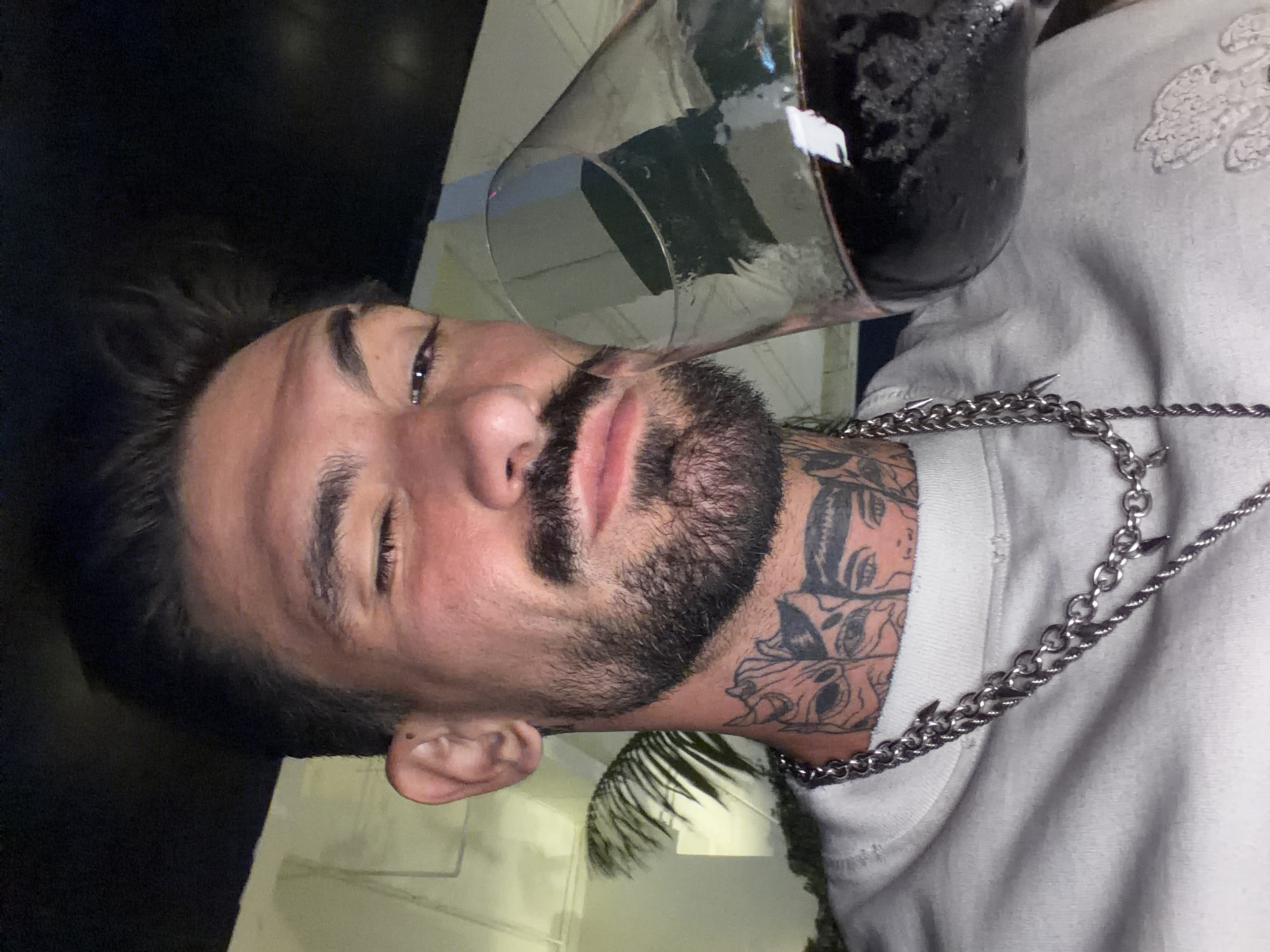 A man is indoors in what appears to be a dimly lit bar or restaurant, holding a glass of dark liquid. He has dark hair, a beard, and tattoos on his neck. He wears a gray sweatshirt and a chain necklace with metal spikes. The location is indicated as Pattaya City, Thailand.