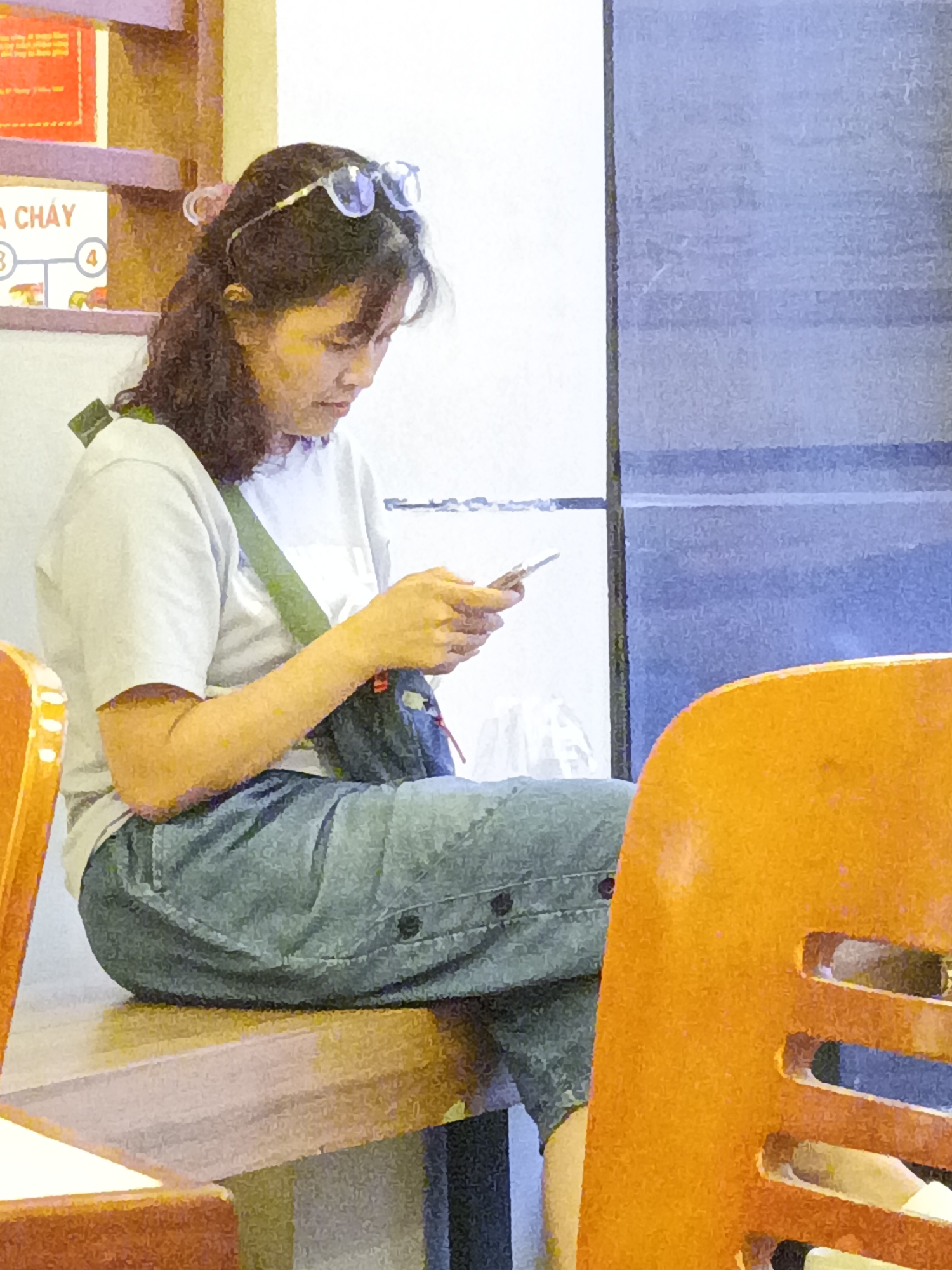 An Asian woman with dark hair pulled back and wearing glasses is sitting on a wooden surface, looking down at her phone. She is wearing a white t-shirt, a green strap across her chest, and loose-fitting denim pants. A dark bag is slung across her body. In the background, a shelf displays a sign with Vietnamese text, including the words "CHAY" and the numbers "3" and "4". An orange chair is visible in the foreground on the right side of the image. The overall setting appears to be indoors, possibly a cafe or restaurant in Nha Trang Ward, Vietnam, given the contextual information provided.