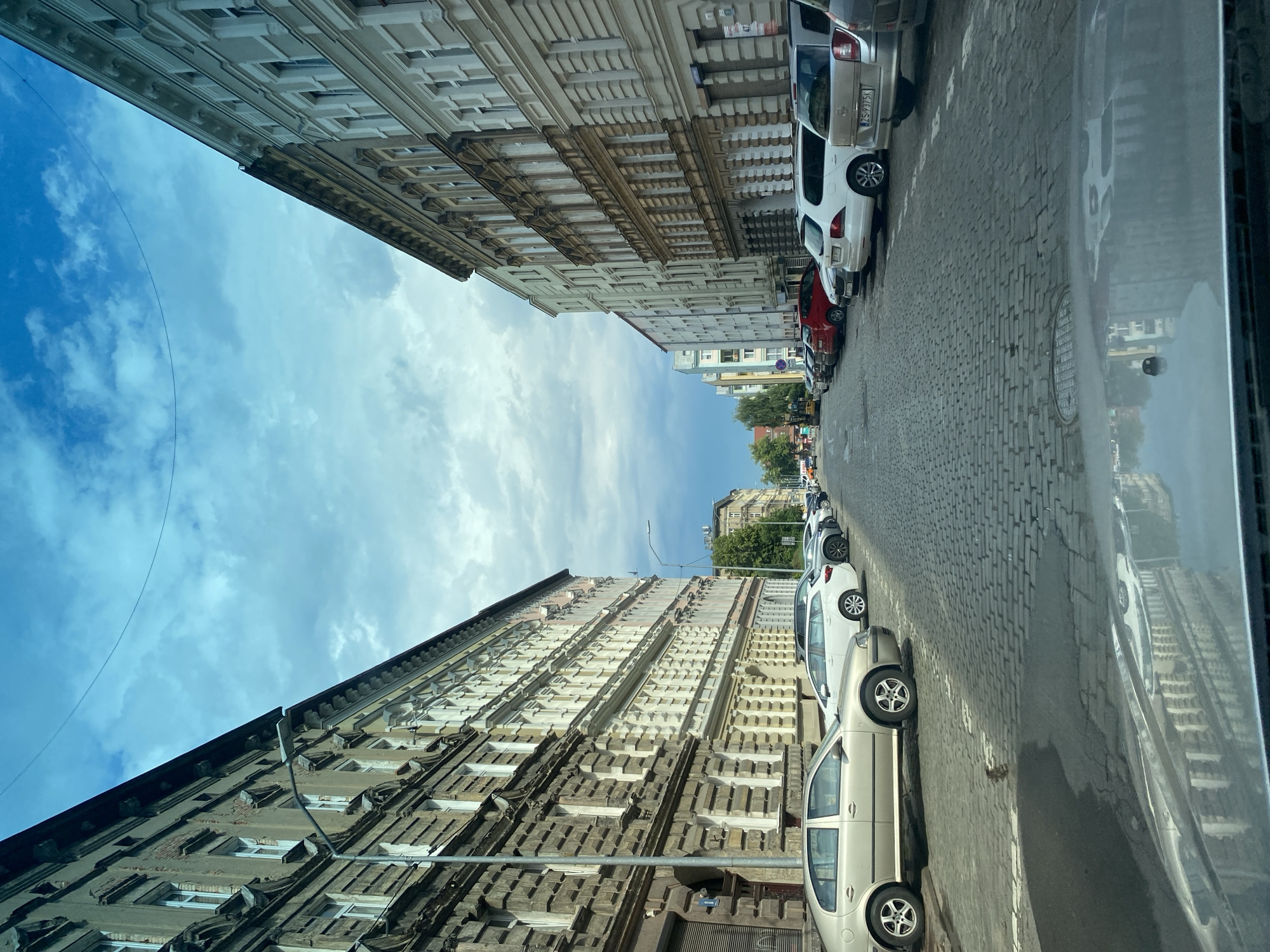 A low-angle, medium shot captures a street scene in Szczecin, Poland, with traditional European architecture lining both sides of a cobblestone road. The street is filled with parked cars, and the sky above is a mix of blue and white clouds.
On the left side of the street, a weathered, multi-story building features a dark facade with ornate detailing around the windows and along the roofline. Some of the brickwork appears damaged, revealing older layers beneath. A street lamp with a modern fixture is positioned in front of this building. To the right of this building, a lighter-colored, also multi-story structure with numerous windows is visible.
On the right side of the street, a series of buildings showcase a lighter, more uniform facade, likely painted. These buildings also have a repeating pattern of windows and decorative elements.
The street itself is paved with cobblestones, appearing wet in some areas, indicating recent rain or a cleaning. Several cars are parked along both sides of the street, including a silver sedan in the foreground on the left, a white station wagon and a silver hatchback on the right. The reflection of the buildings and sky can be seen on the hood of a vehicle in the immediate foreground.
In the distance, the street continues, with more buildings and trees visible. The overall atmosphere suggests an urban environment in a historic European city. The presence of parked cars and the clear visibility of building facades and street paving indicate a street in a residential or mixed-use area.