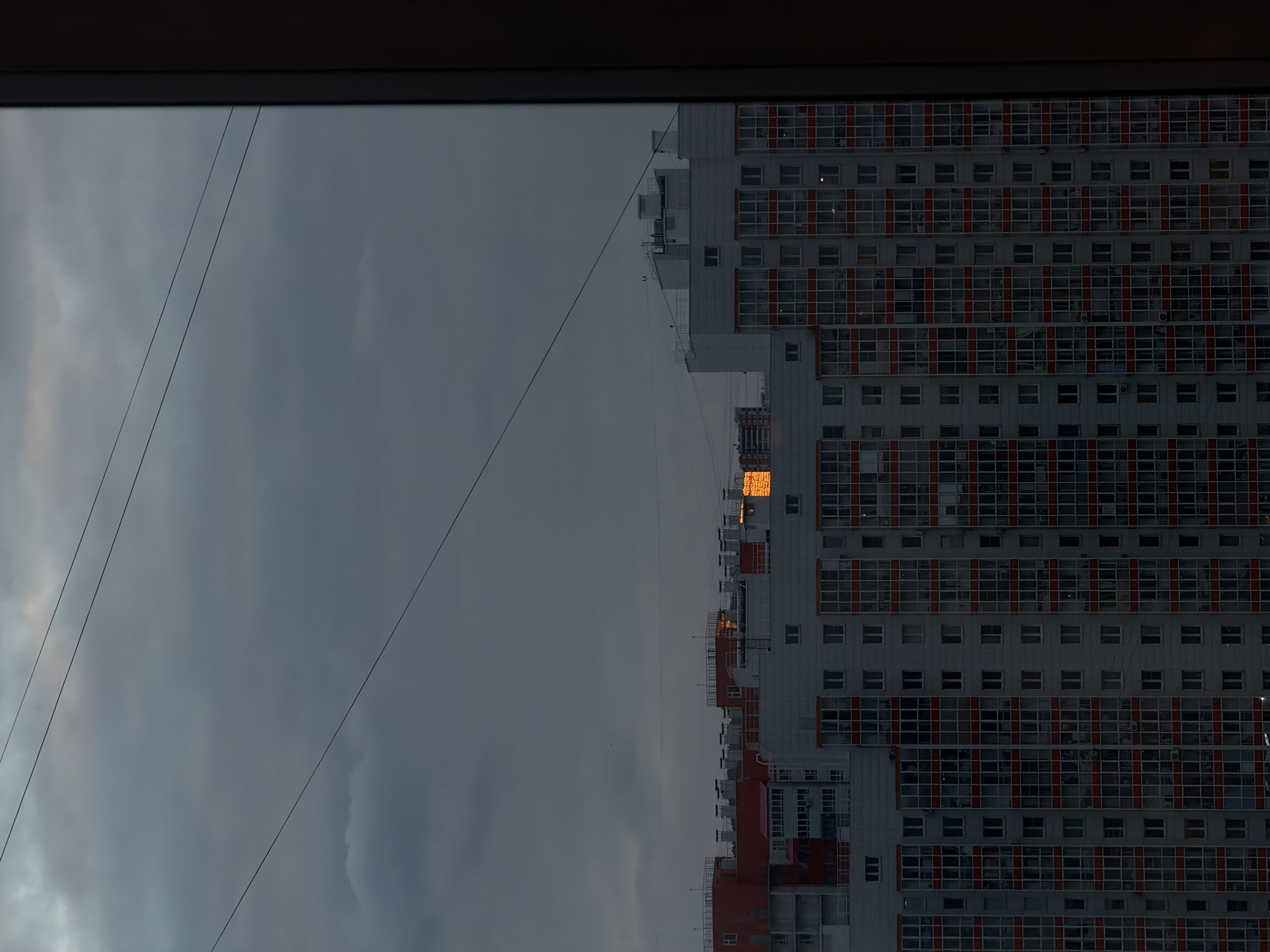 The image is a view from inside a building looking out towards another tall, multi-story residential building in Krasnoyarsk, Russia. The frame of the window is visible at the top and the left side. The building across the street is primarily white and features a grid-like pattern of windows and reddish-orange vertical stripes. Some of the windows reflect a bright, warm light. The sky is overcast and a dull gray, with a few electrical wires stretching across the left side of the view.