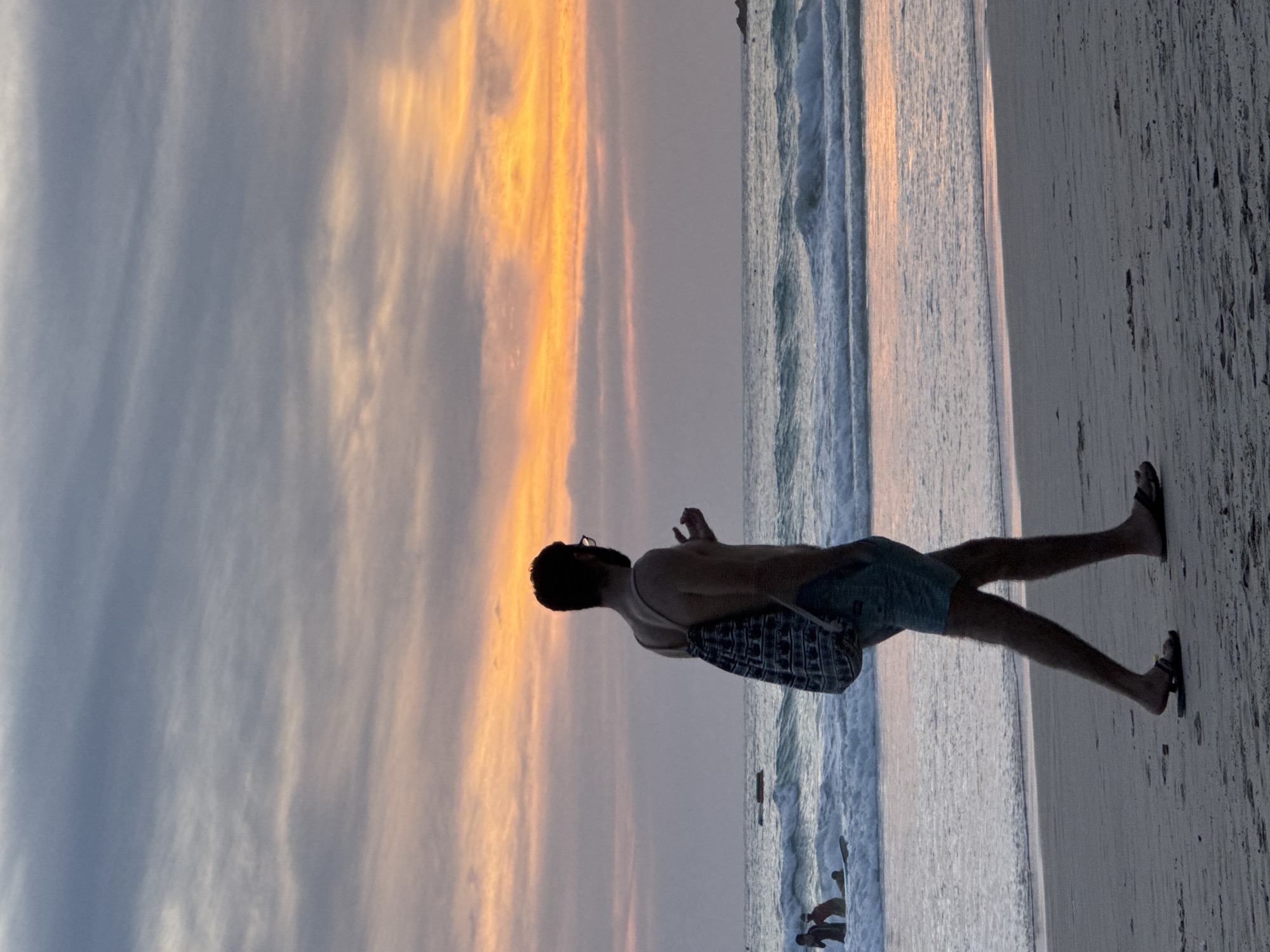 In Karon, Thailand, at sunset, a man walks along a beach. He is silhouetted against the colorful sky, which is painted with hues of orange, pink, and grey. The man is wearing swim trunks and flip-flops, and a patterned bag is slung over his shoulder. He holds his right hand up, fingers slightly splayed, in a gesture that could be conversational or contemplative. In the background, the ocean rolls in with gentle waves, and a few figures are visible in the water, some with a surfboard. The sand is a soft, greyish hue, marked with the footprints of beachgoers and the debris of the tide. The overall scene evokes a sense of calm and reflection as the day transitions into night.