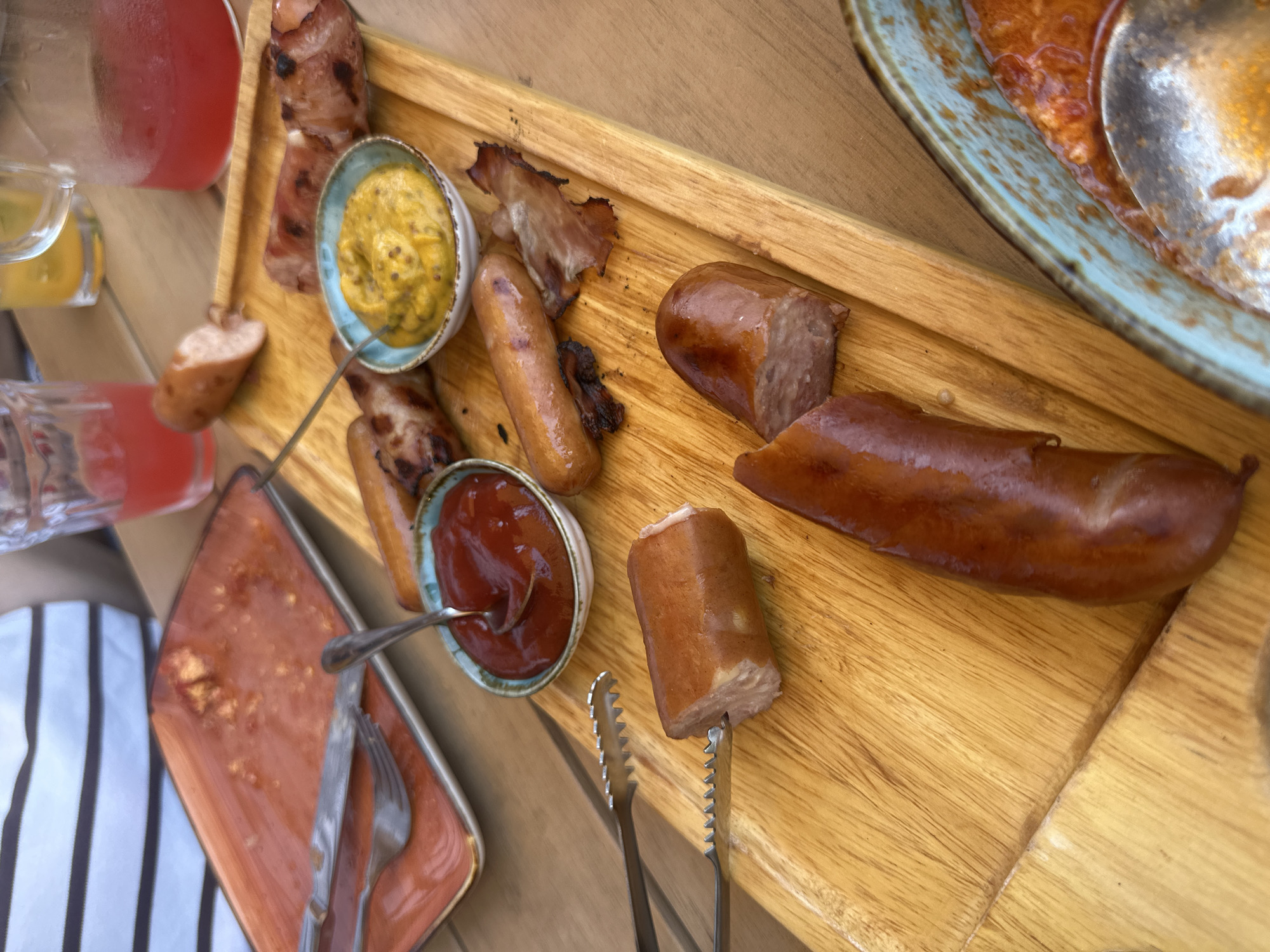 In Byurakan, Armenia, a wooden board filled with sausages and various accompaniments is presented on a table. The board holds several cooked sausages, some whole and some sliced, along with pieces of crispy bacon. Two small bowls are also on the board: one contains a dollop of mustard, and the other holds ketchup. Serving tongs are positioned to pick up a sausage slice. To the left of the board, a partially eaten meal is visible on an orange rectangular plate, with a fork and knife resting on it. In the background, a person's arm wearing a horizontally striped shirt is visible, along with glasses and pitchers of red and yellow beverages. A blue-rimmed bowl containing a red sauce with a spoon is also partially visible in the lower right corner of the image. The overall scene depicts a casual meal, likely outdoors, given the wooden table and natural lighting.