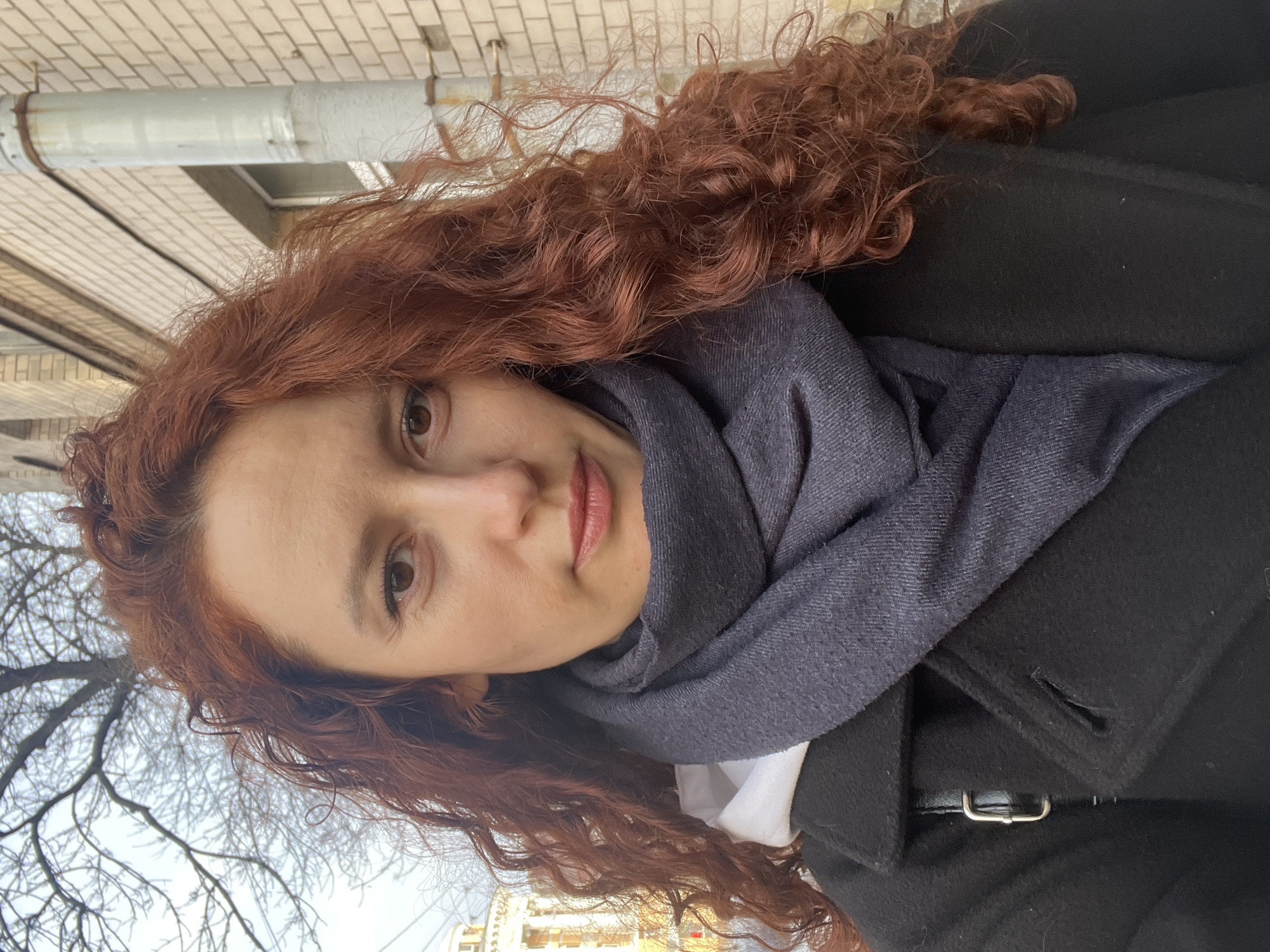A woman with long, curly, auburn hair is wearing a dark coat and a gray scarf. She is standing outside on a street. In the background are buildings and trees, consistent with an urban environment. The architecture in the background appears to resemble buildings in Saint Petersburg, Russia. A drainpipe is visible on the upper left. The woman is looking at the camera.