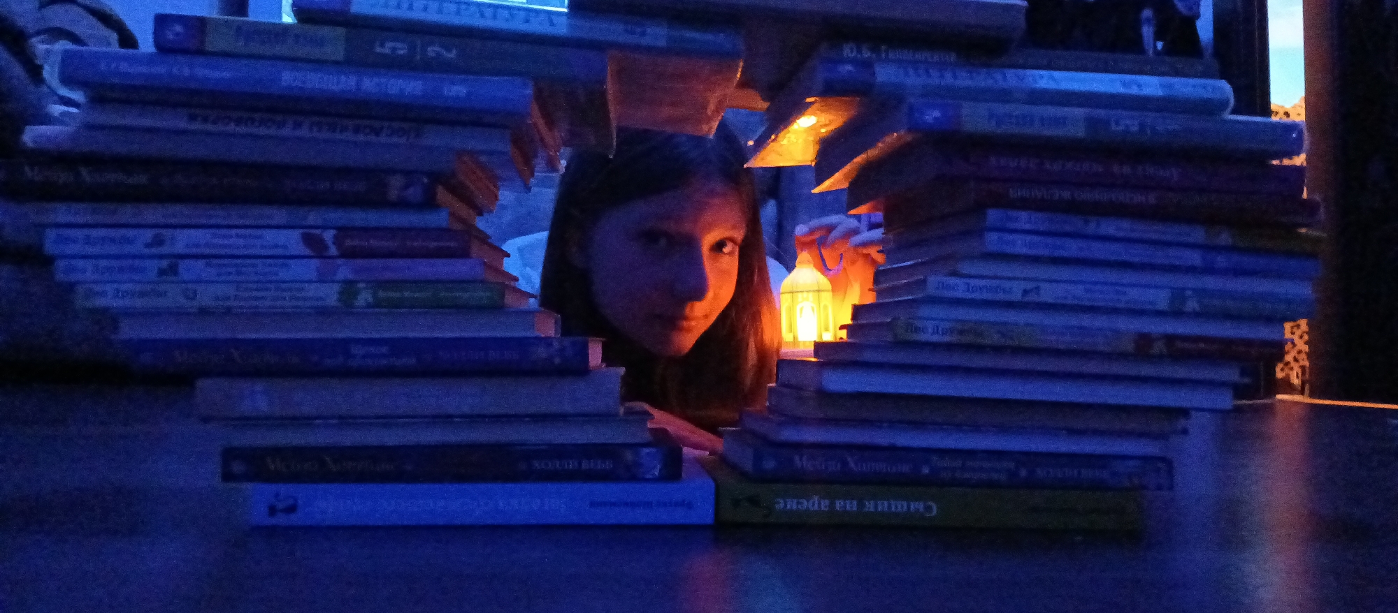 The image features a young person, appearing to be a girl, peeking through a creative structure built from stacks of books. The scene is set indoors, likely in a room during the evening or night, indicated by the dim, atmospheric lighting. A cool blue ambient light pervades the background and the majority of the book spines, contrasting sharply with the warm, orange glow emanating from a small, ornate lantern placed within the book fort.
The girl's face is illuminated by this warm light, drawing attention to her eyes and a subtle, direct gaze towards the viewer, conveying a sense of curiosity or quiet playfulness. Books are piled high on a flat, dark surface, forming two substantial walls that create a narrow, tunnel-like opening in the center.
The visible text on the book spines, predominantly in Cyrillic script, includes titles such as "Melia Хаттоне" (Melia Hutton), "Сыщик на арене" (Detective in the arena/Sleuth in the arena), and "Лес Друзаба" (Druzhba Forest). Other partial texts like "ВОЕВЕЩАЯ ИСТОРИЕ", "SOUDE", "VALAI", and "НО ЖЕЛАНИЯ" are also discernible. The overall composition creates an intimate and imaginative setting, suggesting a moment of escape or quiet enjoyment within a personal sanctuary of books.