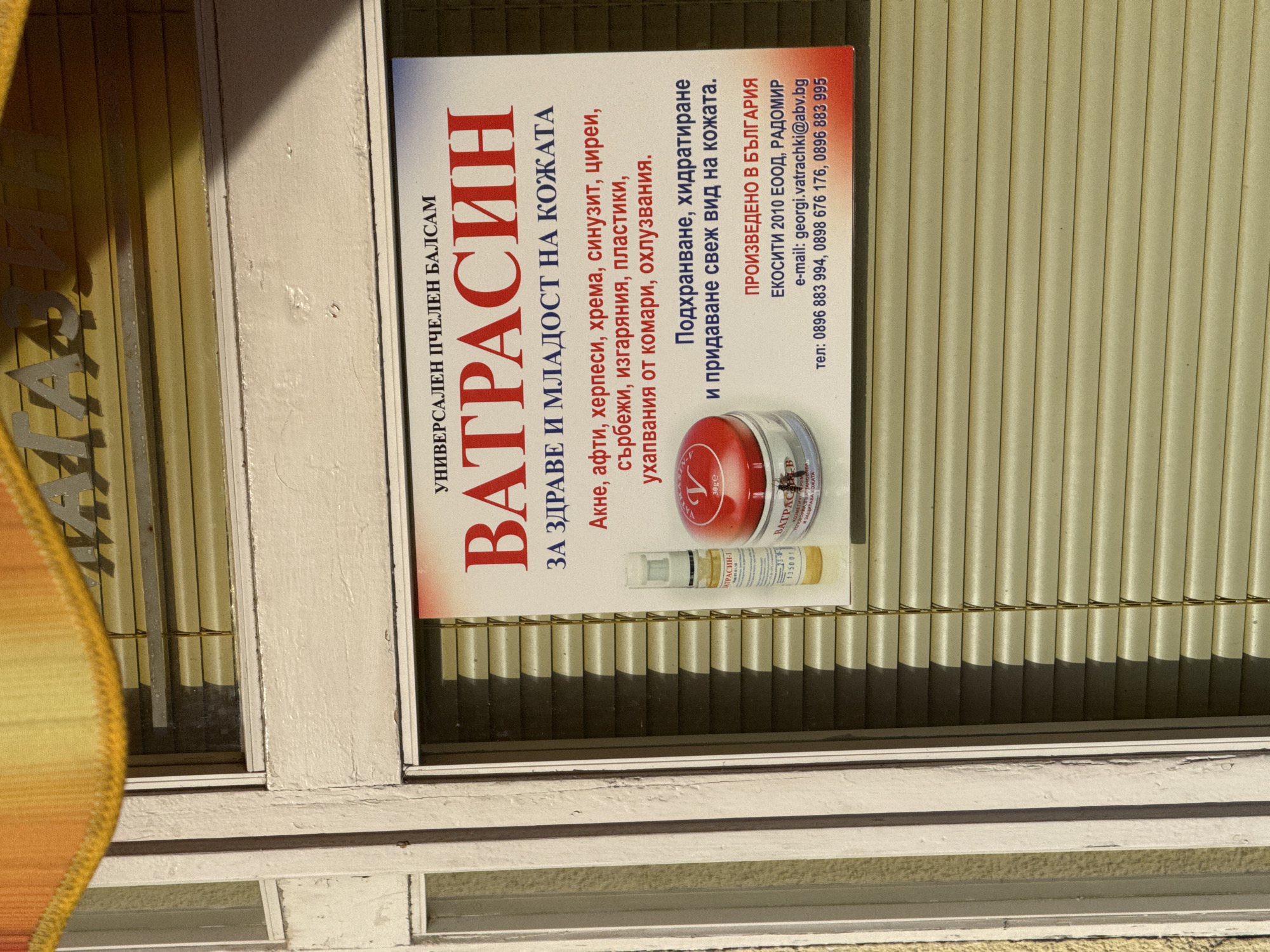 A promotional poster for "Vatrasin" skin balm is mounted on a window frame with slatted blinds visible through the window. The poster is red and white and written in Bulgarian. It lists benefits such as treating acne, herpes, sinusitis, burns, and insect bites. It claims to nourish, hydrate, and give the skin a fresh look. The poster indicates that the balm is produced in Bulgaria by Ecositi 2010 EOOD in Radomir, including email and phone numbers. The window frame is painted white. The location (Lovech, Bulgaria) is not visually discernible.