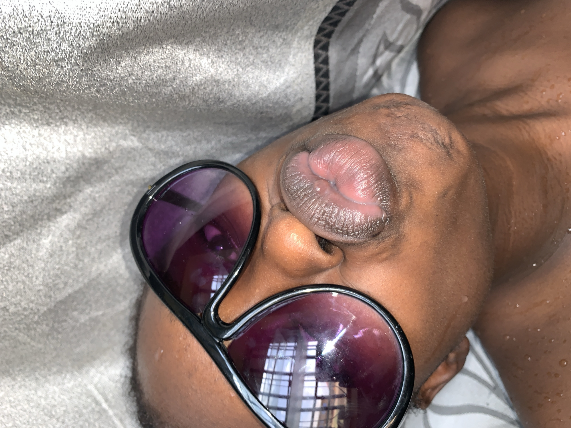 The image shows a person lying down with sunglasses on, viewed from an extreme close-up angle. The person's lips and nose are prominent in the foreground. The sunglasses are black framed with purple-tinted lenses. The background is a light gray fabric. The person's skin is wet. The reflection in the sunglasses' lenses indicates a window with multiple panes. There is no information to determine location in Rafin Par, Nigeria.