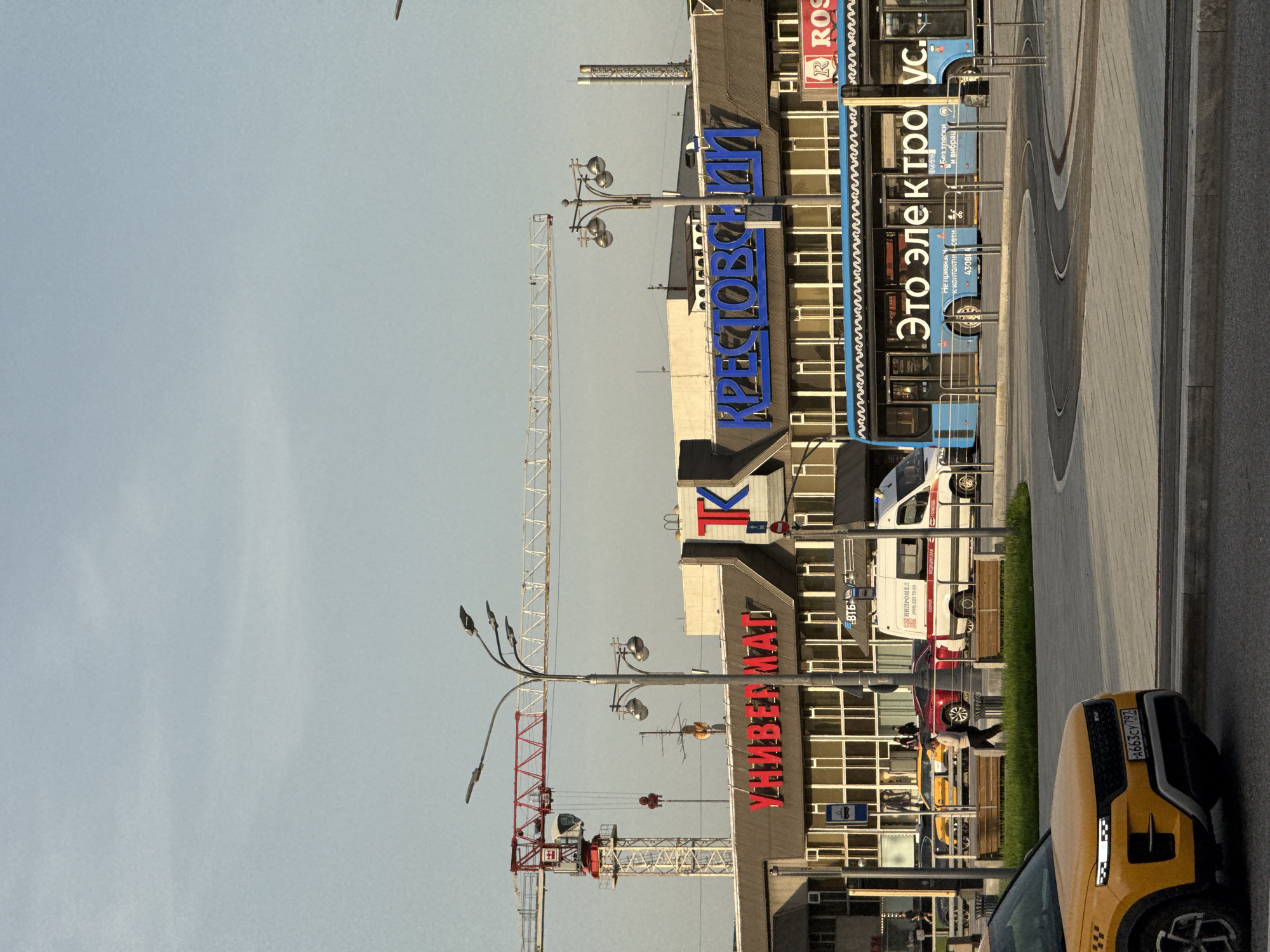 The image depicts a street scene in Moscow, Russia, featuring a shopping center or department store. Several signs are visible on the building, including "Универмаг" (Department Store) in red letters, "ТК" (Shopping Center) with a red and blue logo, and "Крестовский" in blue. There's also a sign advertising "Это электроус" (This is electric us), which seems to be related to electric buses.
A yellow taxi is parked on the right side of the image, and a few people are walking nearby. Cranes are visible in the background, suggesting construction or development in the area. A bus stop sign is also visible.