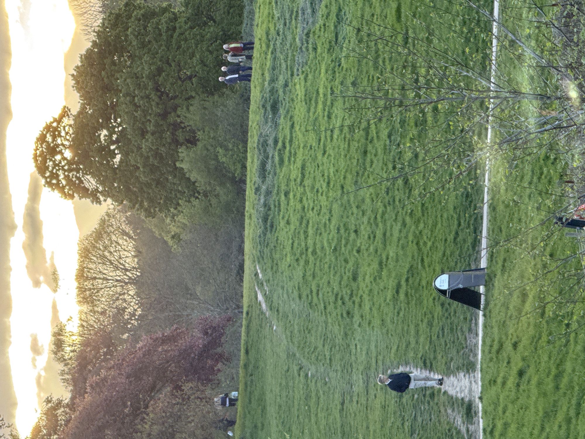 The image depicts a grassy park scene in London, United Kingdom, likely taken during sunset or early evening. The park features a wide expanse of green grass with visible pathways and a number of trees scattered around the landscape. There are people present at different locations; one individual is seen walking along a path, while a group of people is standing at the top of a hill. There is also what appears to be a bench or picnic table near the path. The sky is filled with clouds, and the warm lighting suggests the setting sun.