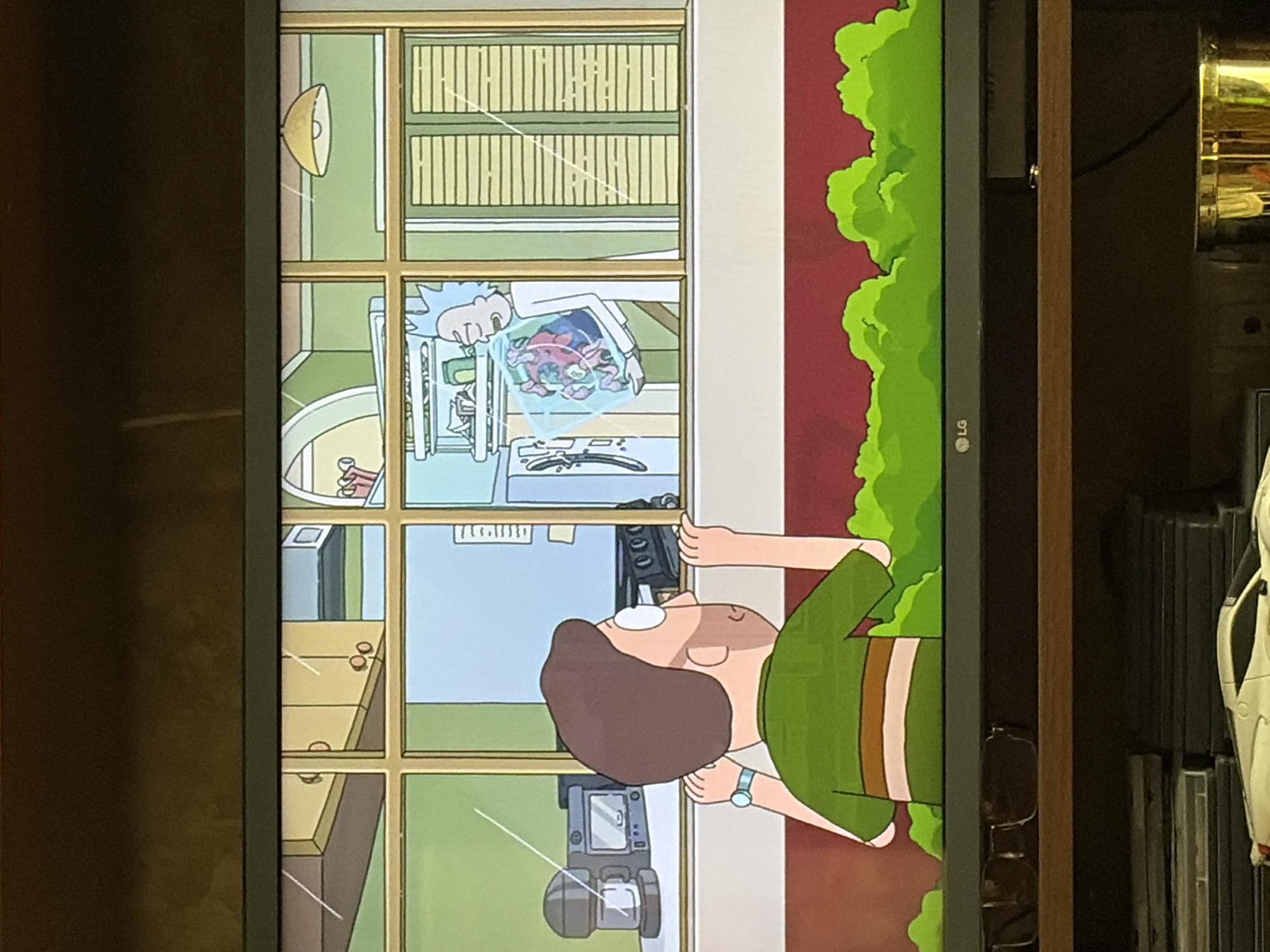 The image shows a television screen displaying an animated scene from the show "Rick and Morty." On the left side of the screen, a character resembling Morty Smith is looking through a window. He is wearing a green shirt with horizontal stripes and a watch on his left wrist. Inside the house, visible through the window, are kitchen appliances, including a microwave and a stove. On the right side of the screen, Rick Sanchez is standing inside the house, holding a clear container with a pink, alien-like creature inside. The background shows a green wall, blinds, and a light fixture. The television itself is black and rests on a wooden shelf. Below the shelf, there are stacks of items, including what appears to be DVD cases and a white toy car.