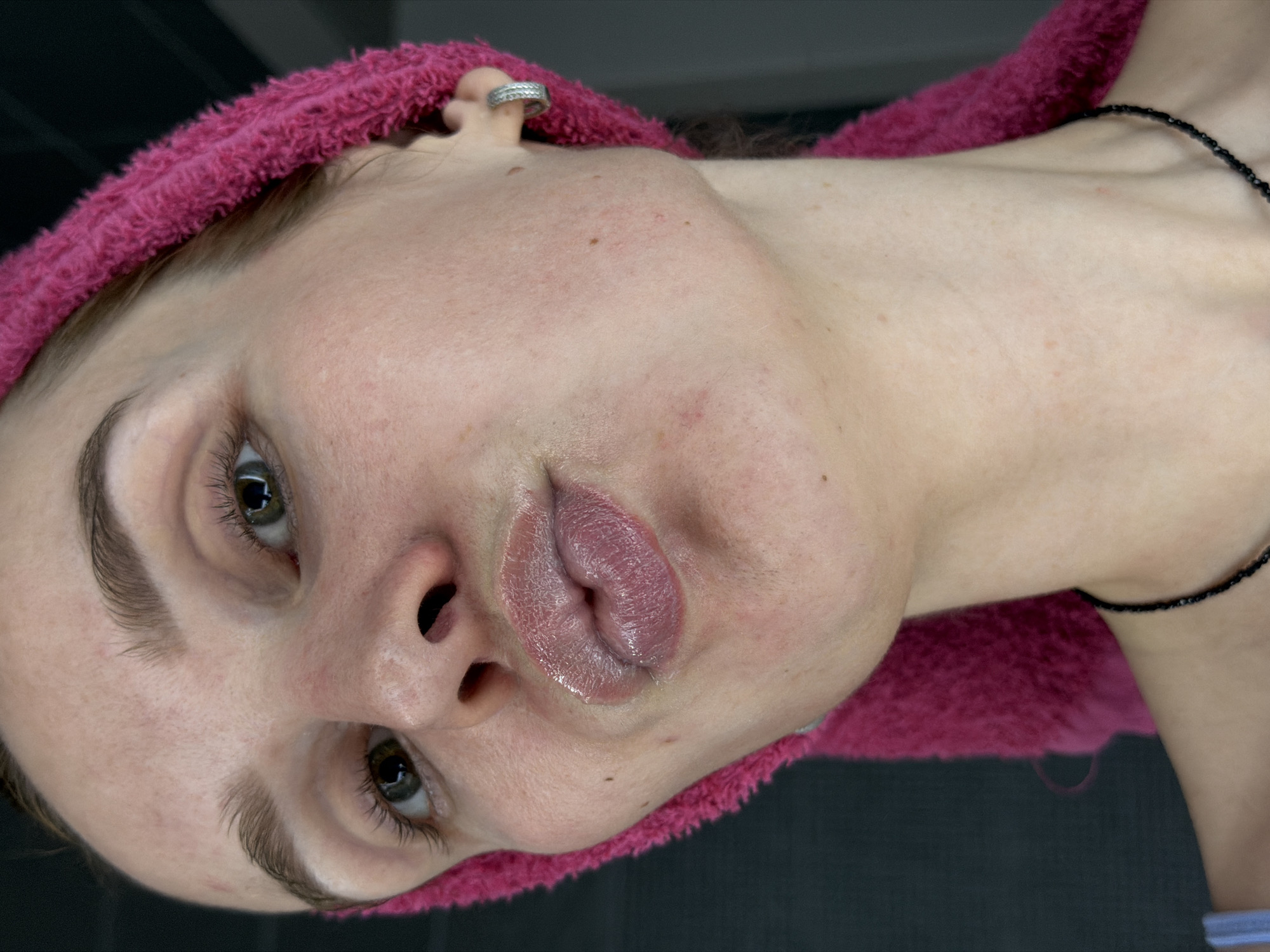 A close-up image shows a fair-skinned woman with her head tilted. A pink towel wraps around her hair. She has light brown eyebrows and green eyes. Her lips appear shiny and are slightly puckered. A silver earring is visible in her left ear. She wears a black beaded necklace. The background is a dark gray. The location is stated as Chaikovsky, Russia, but there are no features in the image to confirm this.