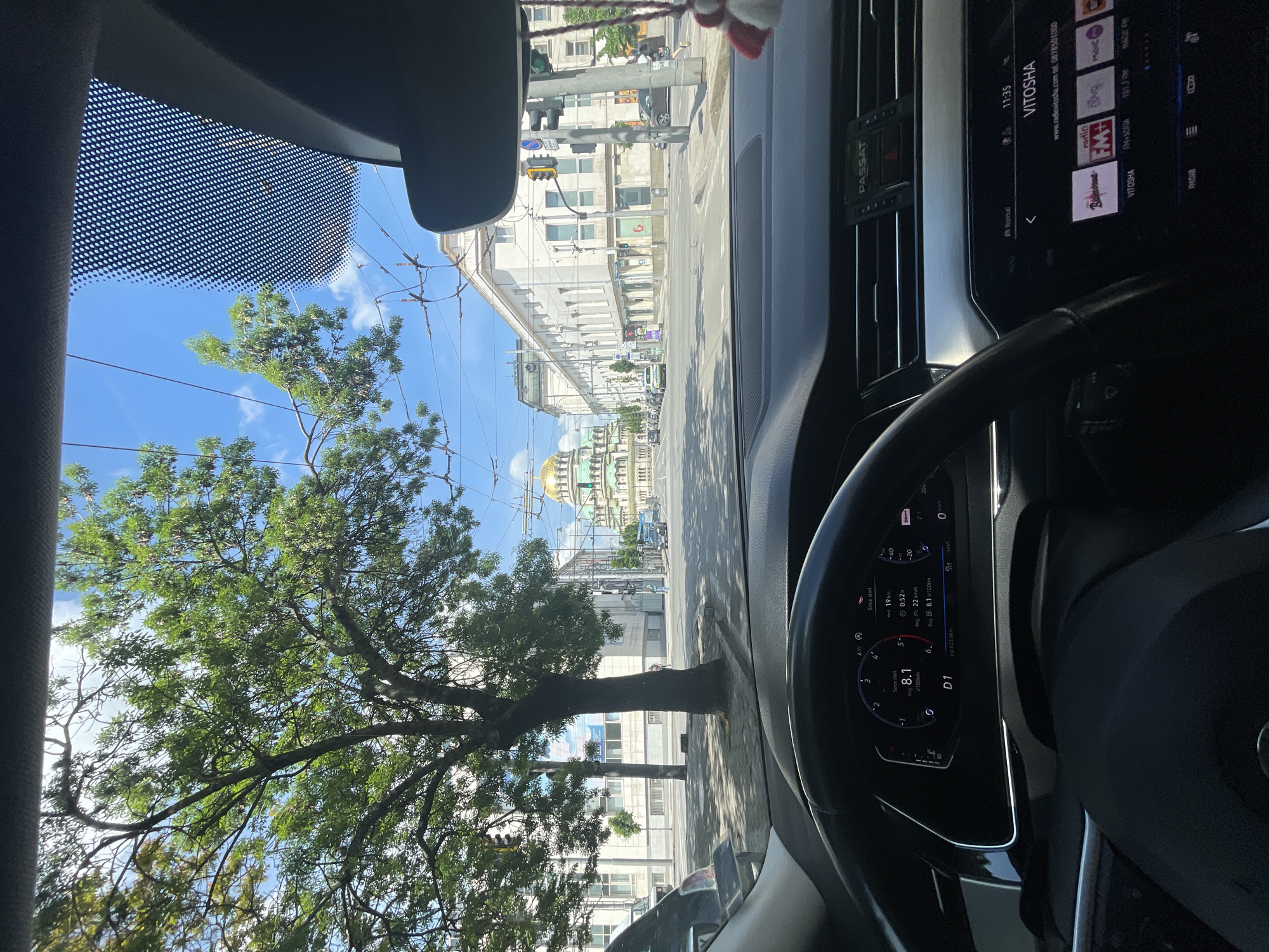 The image is taken from inside a car, likely a Volkswagen Passat, showing the driver's perspective. The car is moving on a street in Sofia, Bulgaria, with a clear view of the Alexander Nevsky Cathedral in the distance, recognizable by its golden dome. A tree partially obstructs the view on the left side of the frame. The car's dashboard is visible, displaying the radio interface tuned to the "VITOSHA" station, and a digital display showing driving statistics like average consumption. The weather appears to be sunny. Other buildings and traffic lights line the street.