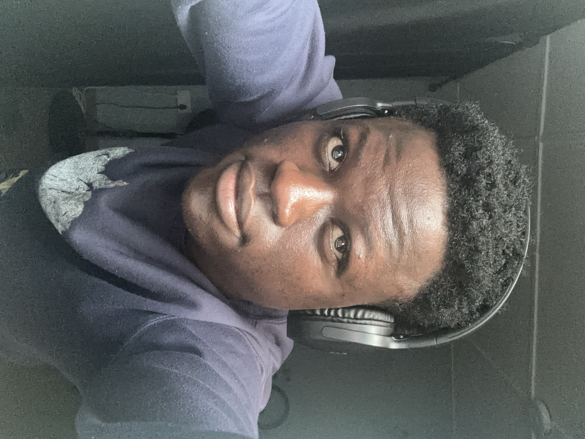 The image is a close-up, frontal selfie of a young Black man wearing over-ear headphones. He has dark, curly hair and is looking directly at the camera with a neutral expression. The man is wearing a dark blue long-sleeved shirt with a light-colored graphic design on the chest. The background is dimly lit and appears to be indoors, with dark walls or drapes. There is a faint image of an electrical outlet and cord visible on the wall behind the man. The lighting in the image suggests it was taken in a low-light environment. The image does not provide any specific visual cues to indicate the location is in Badagry, Nigeria.