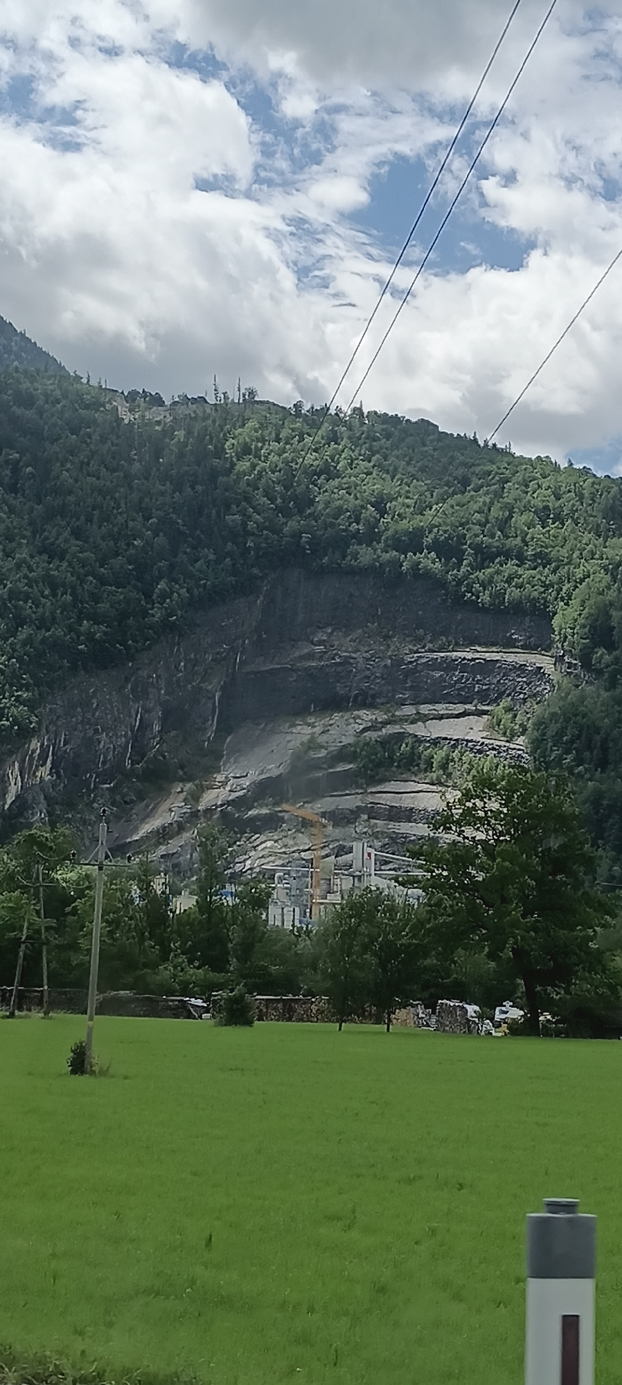 The image depicts a wide shot of a mountainous landscape in Torren, Austria. A steep, forested mountainside dominates the upper half of the frame. A large quarry or mine operation is visible partway up the mountain, characterized by terraced cuts into the rock face and industrial buildings with a yellow crane. The lower portion of the image features a lush green field, with a few trees scattered across it. Several utility poles and lines are visible in the foreground and stretching towards the mountains. The sky is partly cloudy, with patches of blue sky interspersed with white and gray clouds.