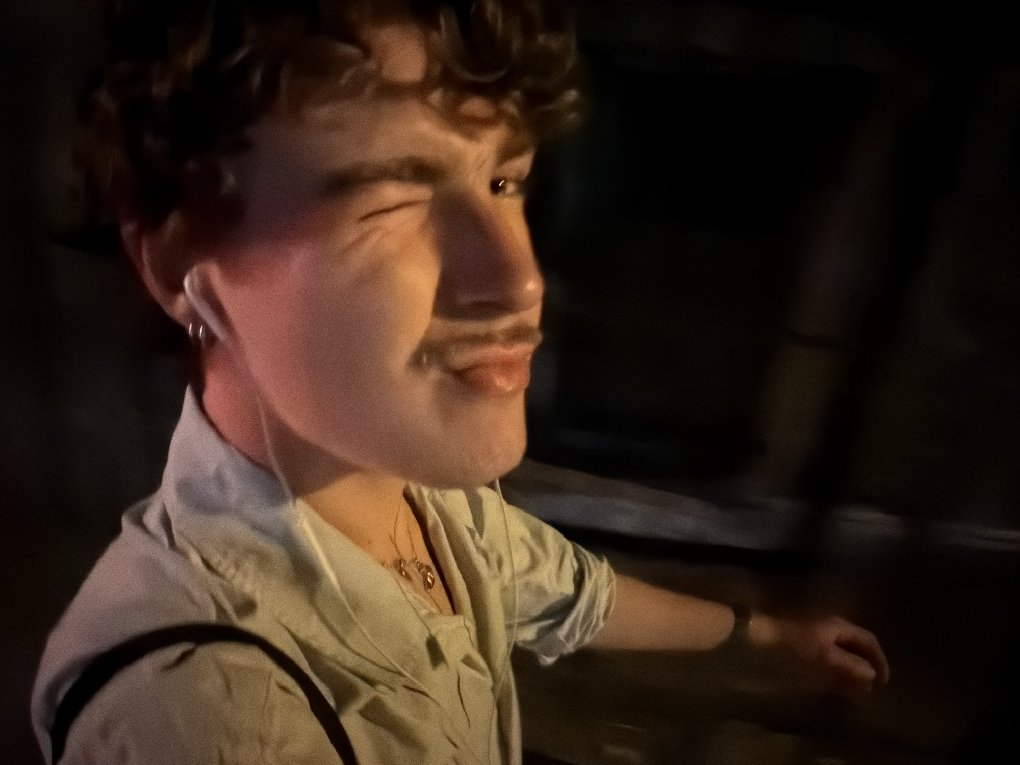 The image shows a close-up, motion-blurred shot of a person winking, with their left eye closed and their right eye open. The person has curly brown hair, a faint mustache, and is wearing a light-colored collared shirt. A white earbud is visible in their left ear, and two gold necklaces are layered around their neck. The lighting is dim with warm tones, suggesting an indoor or nighttime setting. The background is dark and out of focus, offering no specific environmental details. The image does not contain information that would allow for the identification of the location as Vracar Urban Municipality, Serbia.
