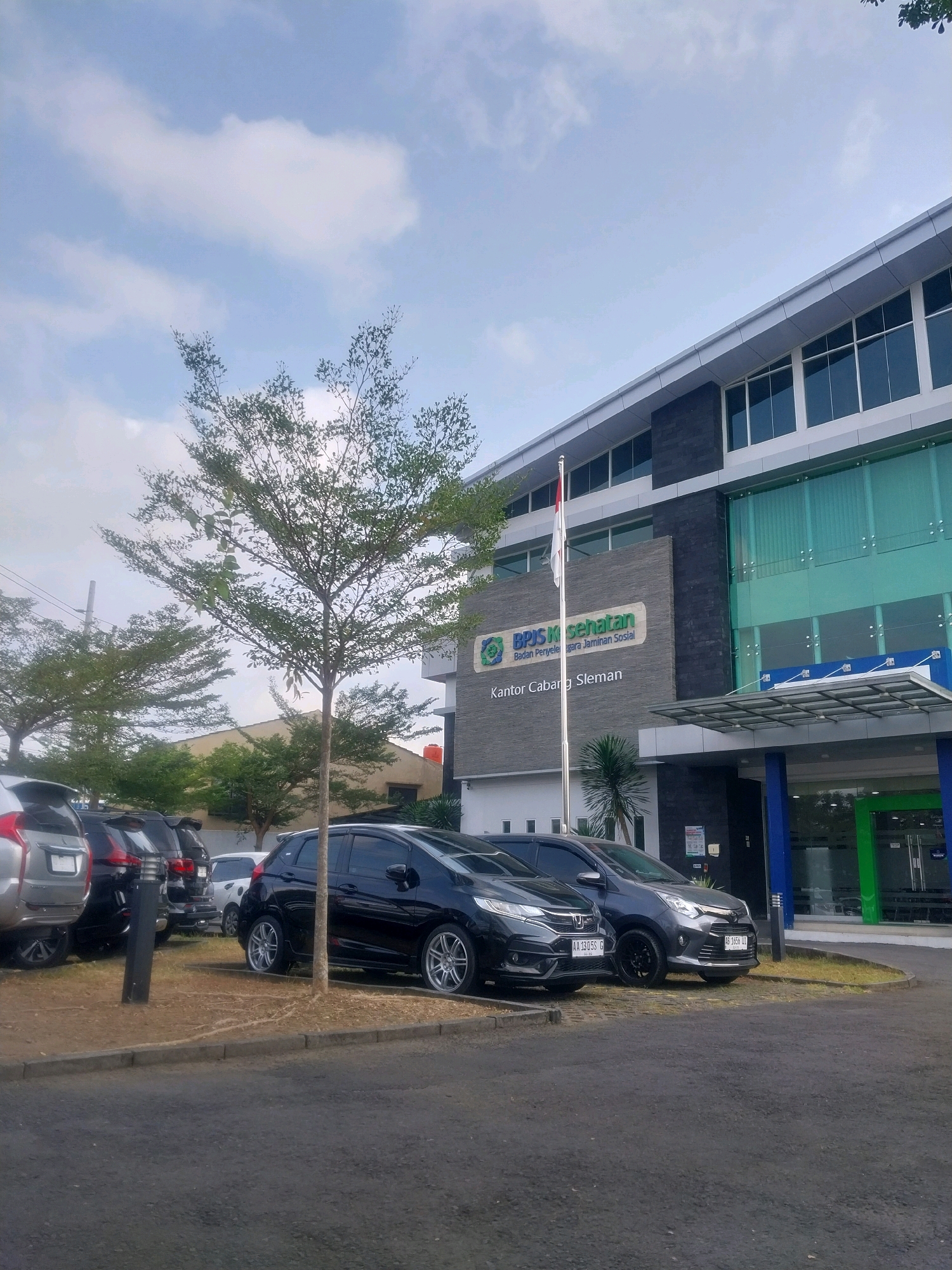 The image captures the exterior of the BPJS Kesehatan Kantor Cabang Sleman in Sleman, Indonesia. The building features a modern design with large glass windows and dark gray stone accents. In front of the building, a parking lot is visible with several cars parked. A flagpole with an Indonesian flag stands prominently near the entrance. The scene is illuminated by natural daylight, with a partly cloudy sky overhead. A slender tree with sparse foliage stands in the foreground, and other trees are visible in the background. The overall atmosphere is calm and functional, depicting a public service building in an urban setting.