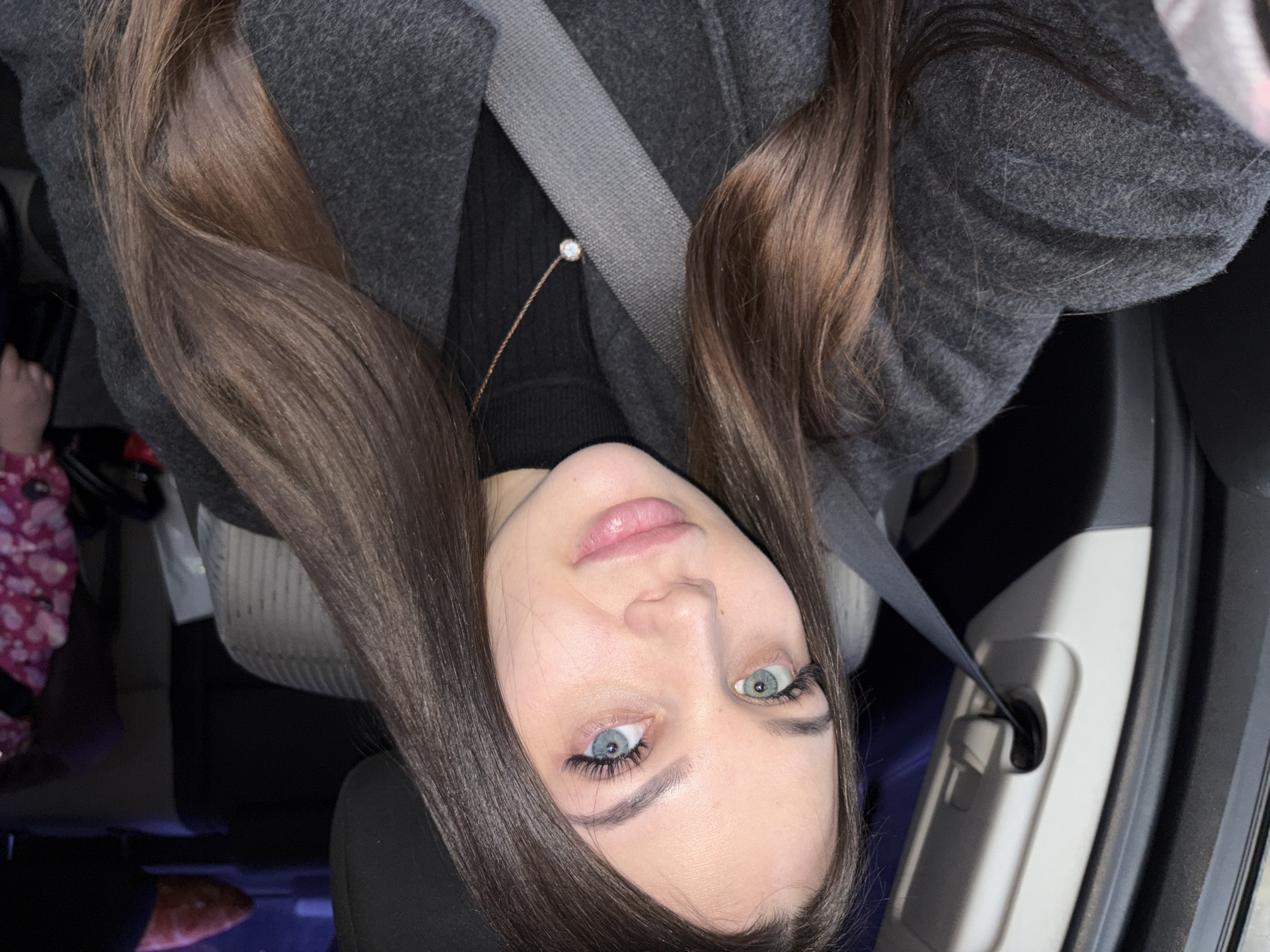 The image shows a woman wearing a dark-colored coat and a seatbelt, suggesting she is in a car. She has long brown hair and distinctive blue and green eyes. A delicate gold necklace with a small pendant is visible. The interior of the car is visible, with seats and door panels showing. A child's foot in a pink floral slipper is visible in the back. The image's location cannot be determined from the provided information.
