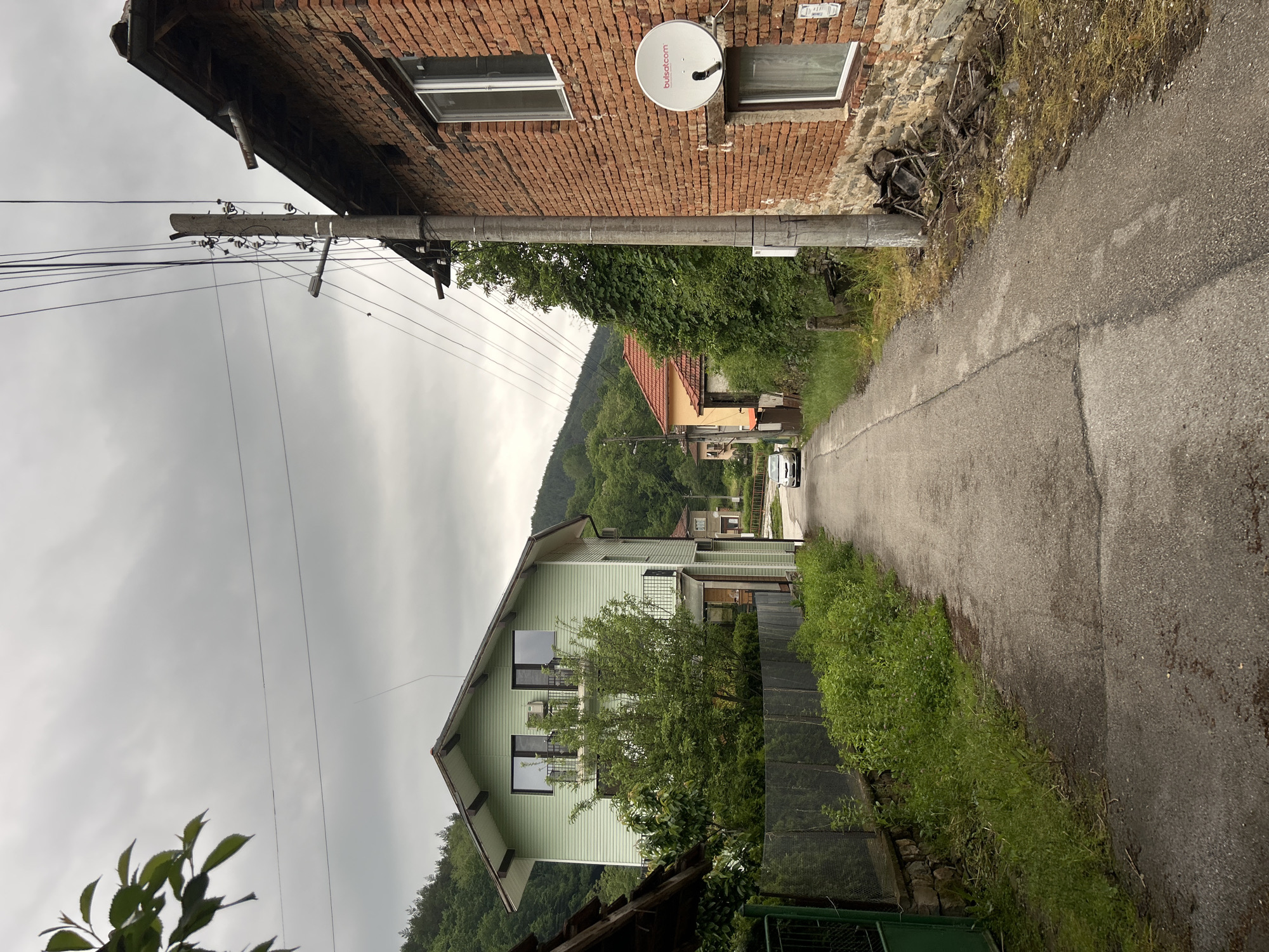 The image depicts a narrow asphalt road running through a rural area. On either side of the road are buildings. To the right is a red brick building with a white satellite dish and two windows. To the left, there is a light green house with a dark roof. A utility pole with numerous wires is positioned near the brick building. In the distance, the road leads towards a small white vehicle and is framed by trees and hills. The sky is overcast. The location is identified as being in an unknown city.