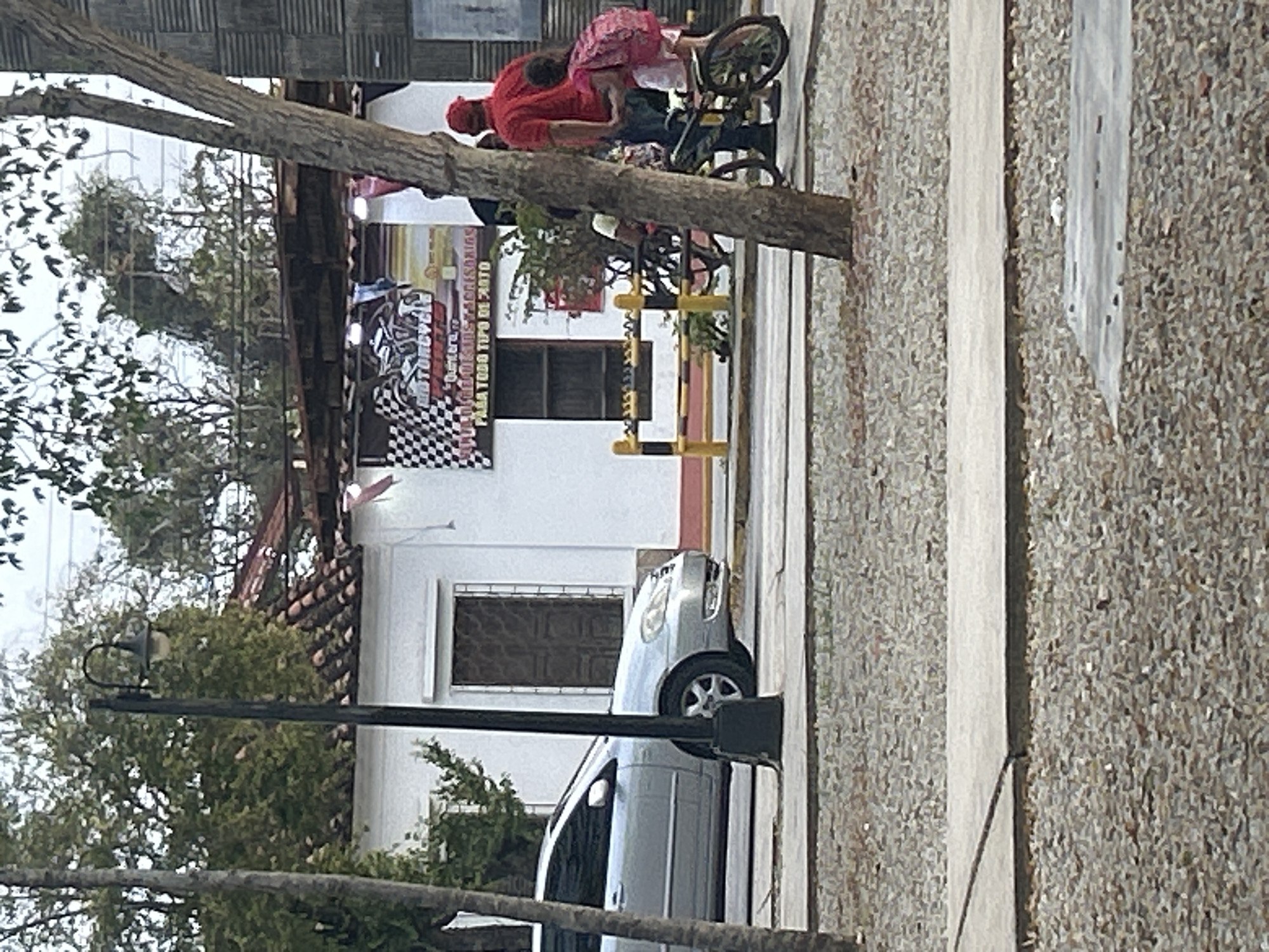In Santa Ana, Venezuela, a silver sedan is parked on a street. A man in a red shirt and cap stands next to a young girl wearing a pink backpack and a white skirt, who is sitting on a bicycle. They are near a building with a white facade and a tiled roof, which has a banner advertising "MOTORCYCLE ACCESORIES". A street lamp is visible to the left of the sedan. The ground is covered in gravel.