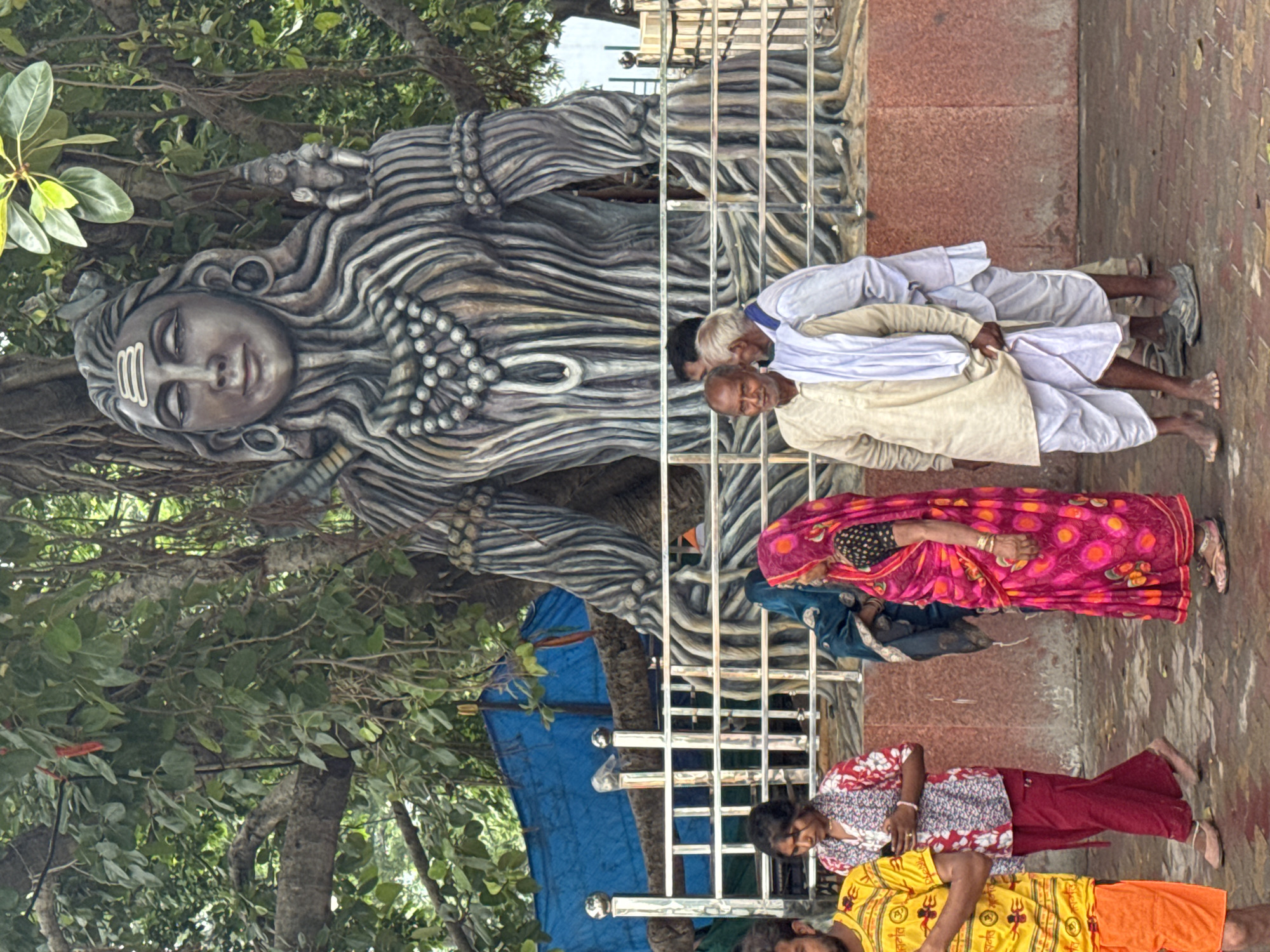 The image depicts a large statue of a Hindu deity, likely Shiva, carved to resemble a tree trunk with intricate details. The statue is situated outdoors, with a lush green canopy of a large tree providing shade and a backdrop. In front of the statue, a metal railing separates it from a paved walkway where several individuals are present.
In the foreground, a group of people are seen. On the left, a man wearing a bright yellow t-shirt and orange shorts is partially visible. To his right, a woman in a vibrant pink and yellow patterned sari is walking, holding a mobile phone in her hand. Next to her, a woman in a blue headscarf and a dark patterned outfit is standing. Further to the right, two men in traditional Indian attire are conversing; one is wearing a light-colored kurta and dhoti, and the other is in a white kurta and dhoti. The ground is paved with reddish-brown bricks, and fallen leaves are scattered on the surface. Behind the railing, a blue tarp is visible, possibly for temporary shelter or covering. The overall scene suggests a public space, likely a temple or a spiritual site in Rishikesh, India, given the prominent religious statue and the attire of the people. The lighting indicates it is daytime.