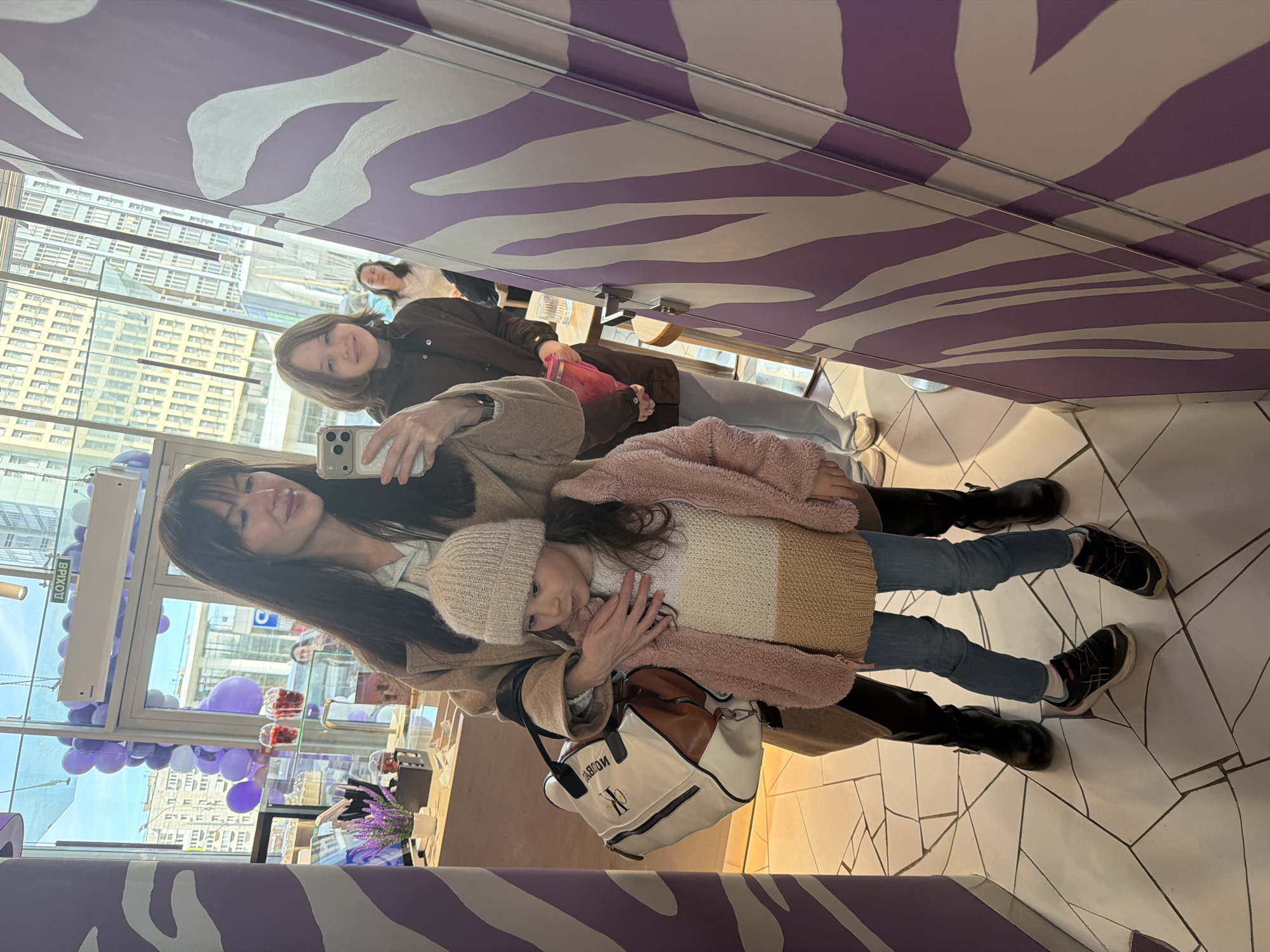 The image is a reflection of several people inside a store, likely in Perm, Russia, as suggested by the window view. The reflection is captured in a mirrored surface with a distinctive zebra-striped purple and white design. In the foreground, a woman with long dark hair is holding a smartphone, apparently taking a selfie. Next to her is a child wearing a knit hat, a striped sweater, and jeans, who appears to be embraced by the woman. Another individual is visible in the background. The store's interior features a display of balloons and possibly merchandise on a counter. Through the window, there is a clear view of a cityscape, suggesting a building with many windows is visible. The floor beneath the mirrored surface consists of white tiles with irregular patterns. The overall scene conveys a casual moment captured in a retail environment.