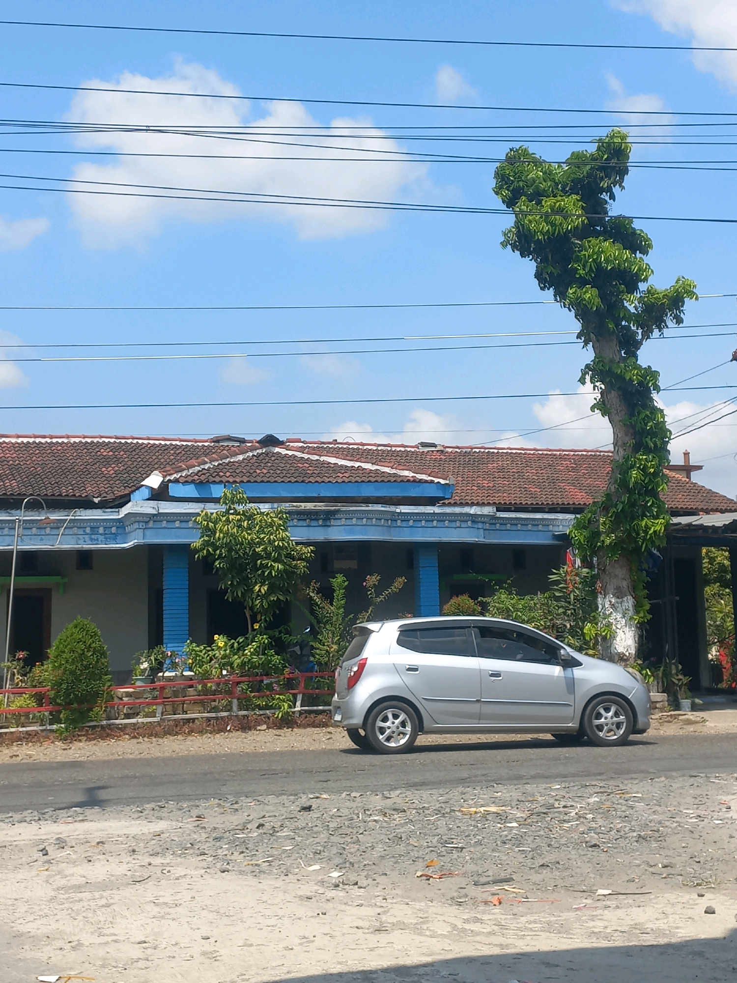 A silver sedan is parked on the side of a paved road in Baturetno, Indonesia. The car is positioned in front of a traditional Indonesian house with a red tiled roof and blue trim. A tall, slender tree with lush green foliage is growing next to the house, its branches reaching upwards and some entwined with vines. Power lines crisscross the bright blue sky, which is dotted with a few white clouds. The foreground of the image shows a rough, gravelly shoulder of the road.