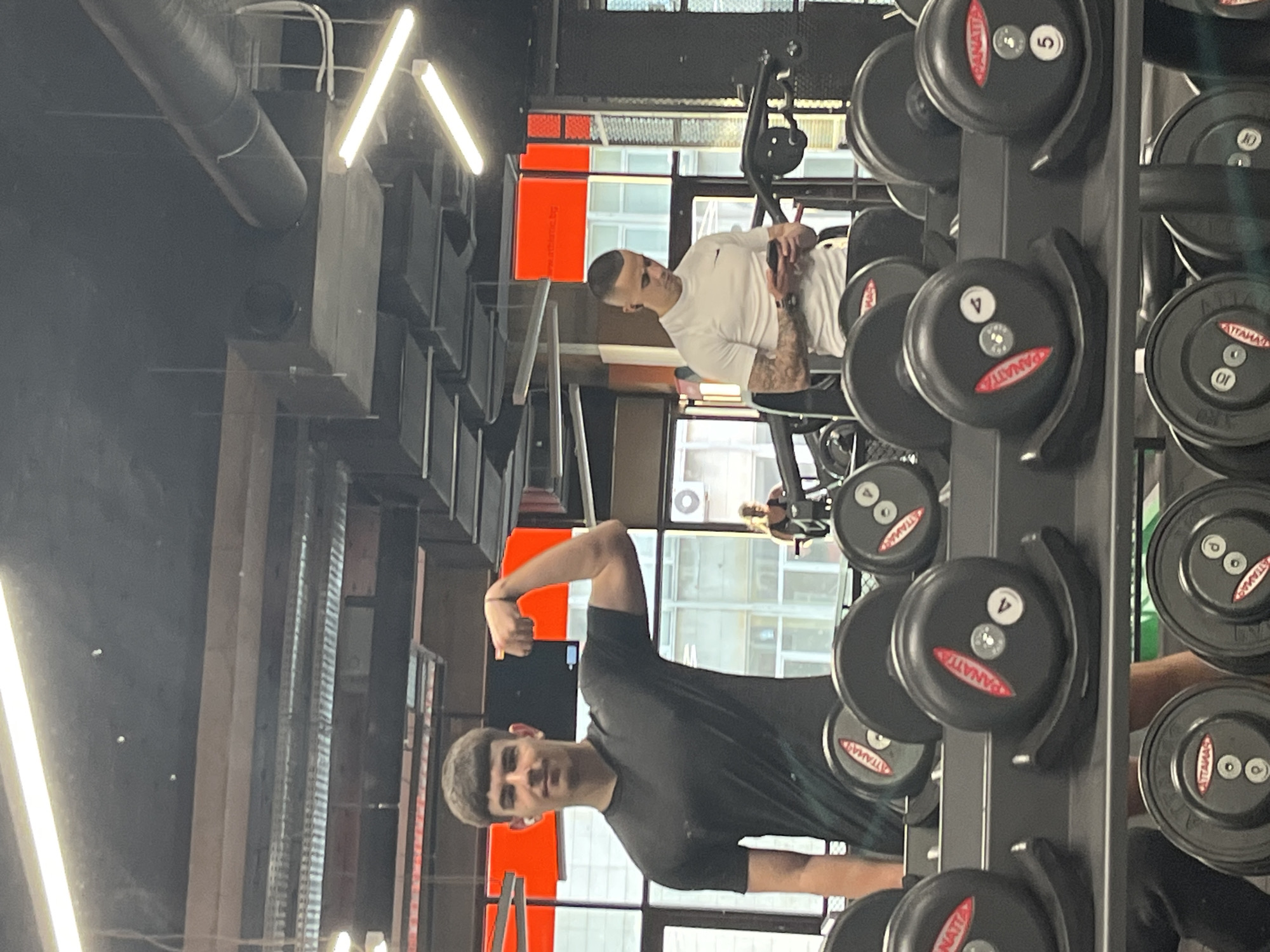 The image depicts the interior of a gym, likely in Plovdiv, Bulgaria, suggested by the presence of “PANATTA” labeled weights (a common gym equipment brand). A young man in a dark T-shirt is flexing his bicep for the camera. In the background, another man wearing a white T-shirt is seated on gym equipment, possibly using a phone. A rack filled with dumbbells of various weights (marked 4, 5, and 10) is prominently featured in the foreground. Large windows are visible behind the people, suggesting an urban setting. The gym has modern lighting and a visible ventilation system on the ceiling. The action appears to be casual, documenting a gym workout session.