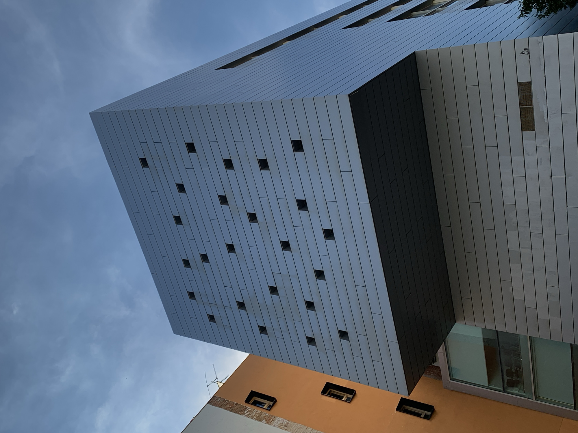 A low-angle shot captures the angular architectural features of a building in Barcelona, Spain. The building showcases a combination of vertical lines and cube-shaped elements. Some sections of the building have a grid of small, dark windows that dot its surface. The sky, filled with blue and grey hues, contrasts with the modern structure. An adjacent orange-colored building and a few hints of greenery can be seen in the foreground.