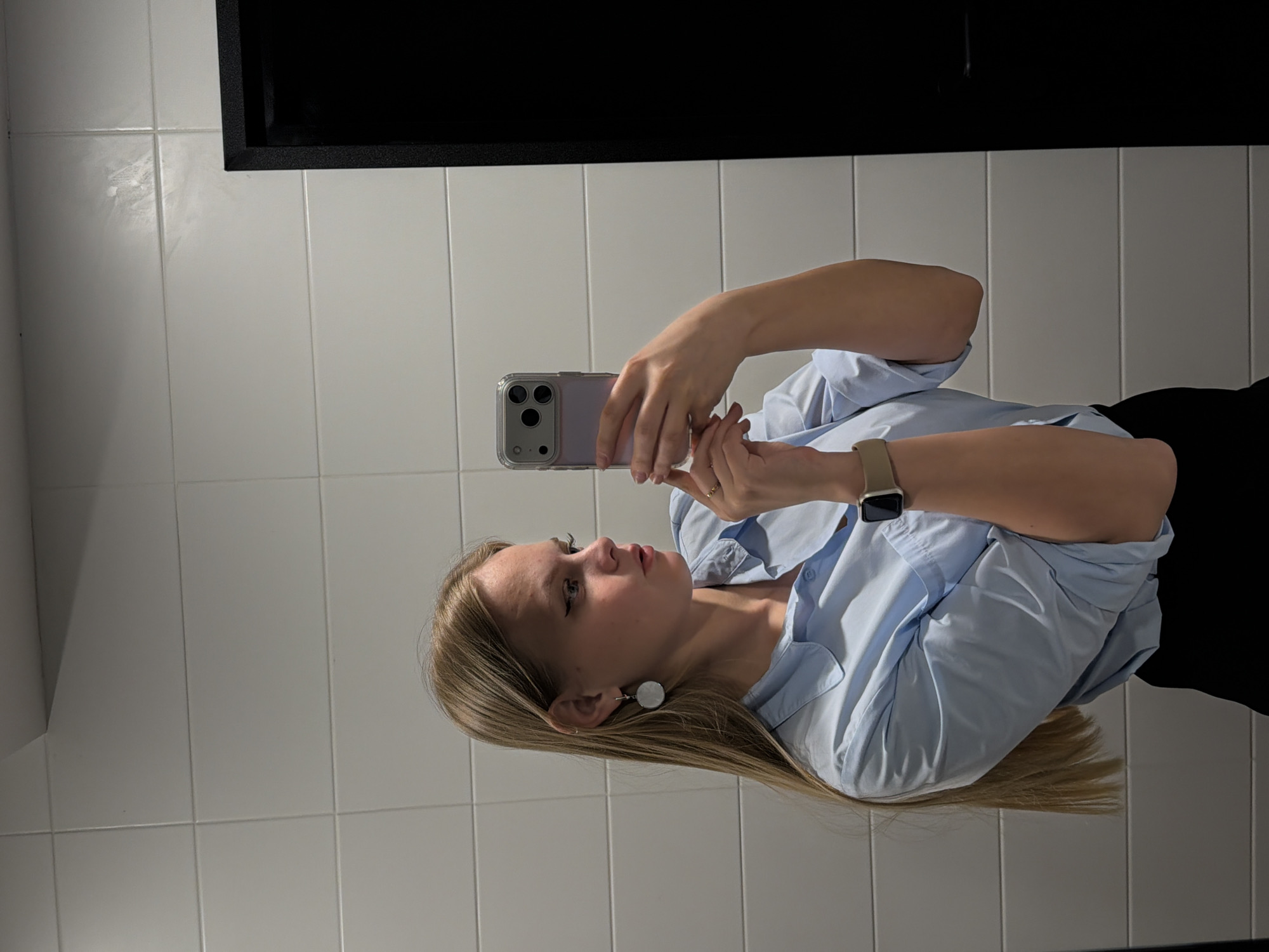 A woman is taking a selfie in a bathroom. She is wearing a light blue button-down shirt, a watch, and earrings. The background consists of white tiles and a black rectangular object, possibly a mirror frame. The location is not identifiable as Izhevsk, Russia, based on the visual information provided.