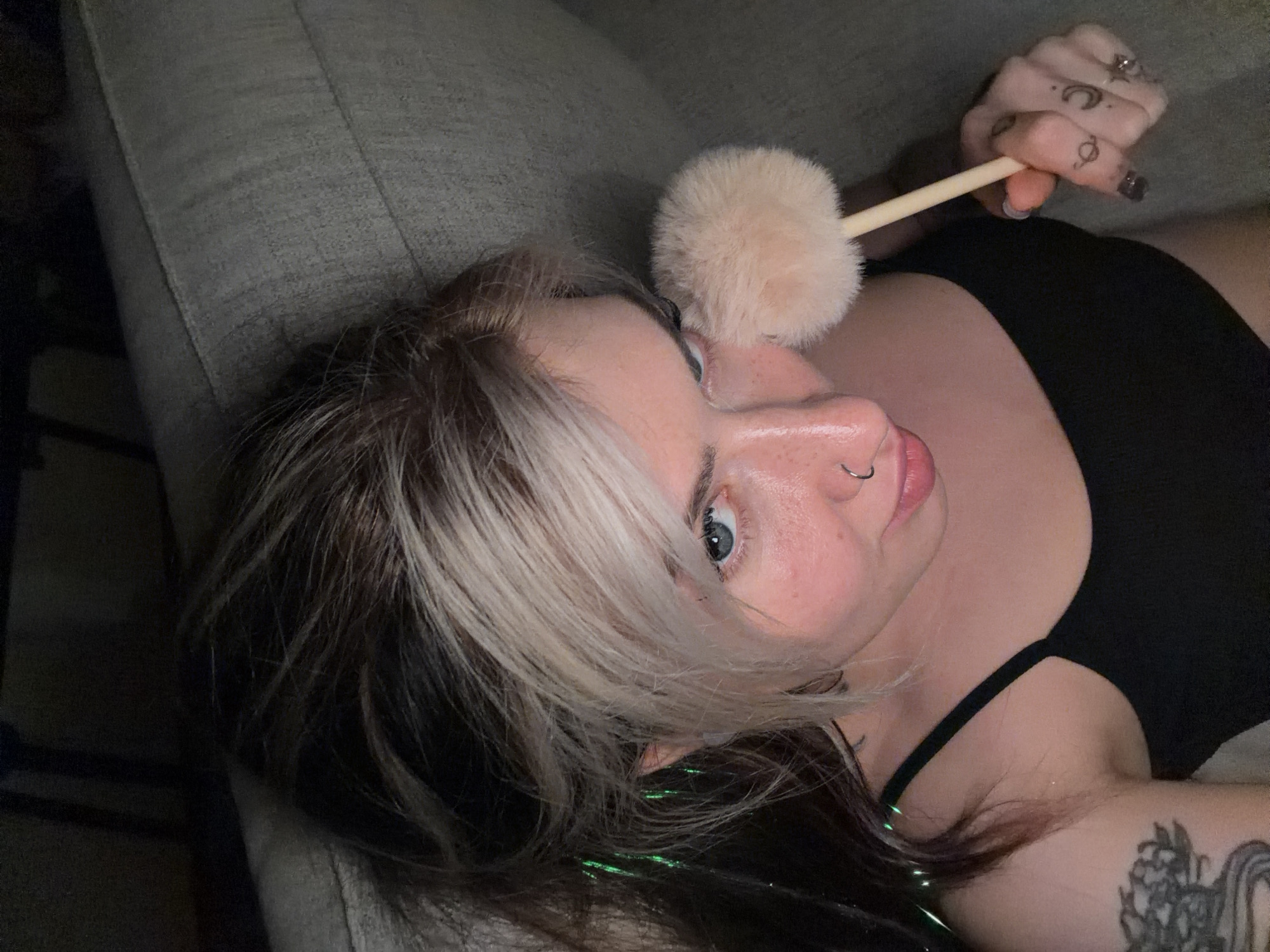 A woman is lying on a gray couch and holding a beige powder brush near her face. She has dyed blond and dark hair and blue eyes. She is wearing a black tank top and has tattoos on her fingers and arm. There are also green tinsel extensions in her hair. There are no indicators of the city of Budva, Montenegro.