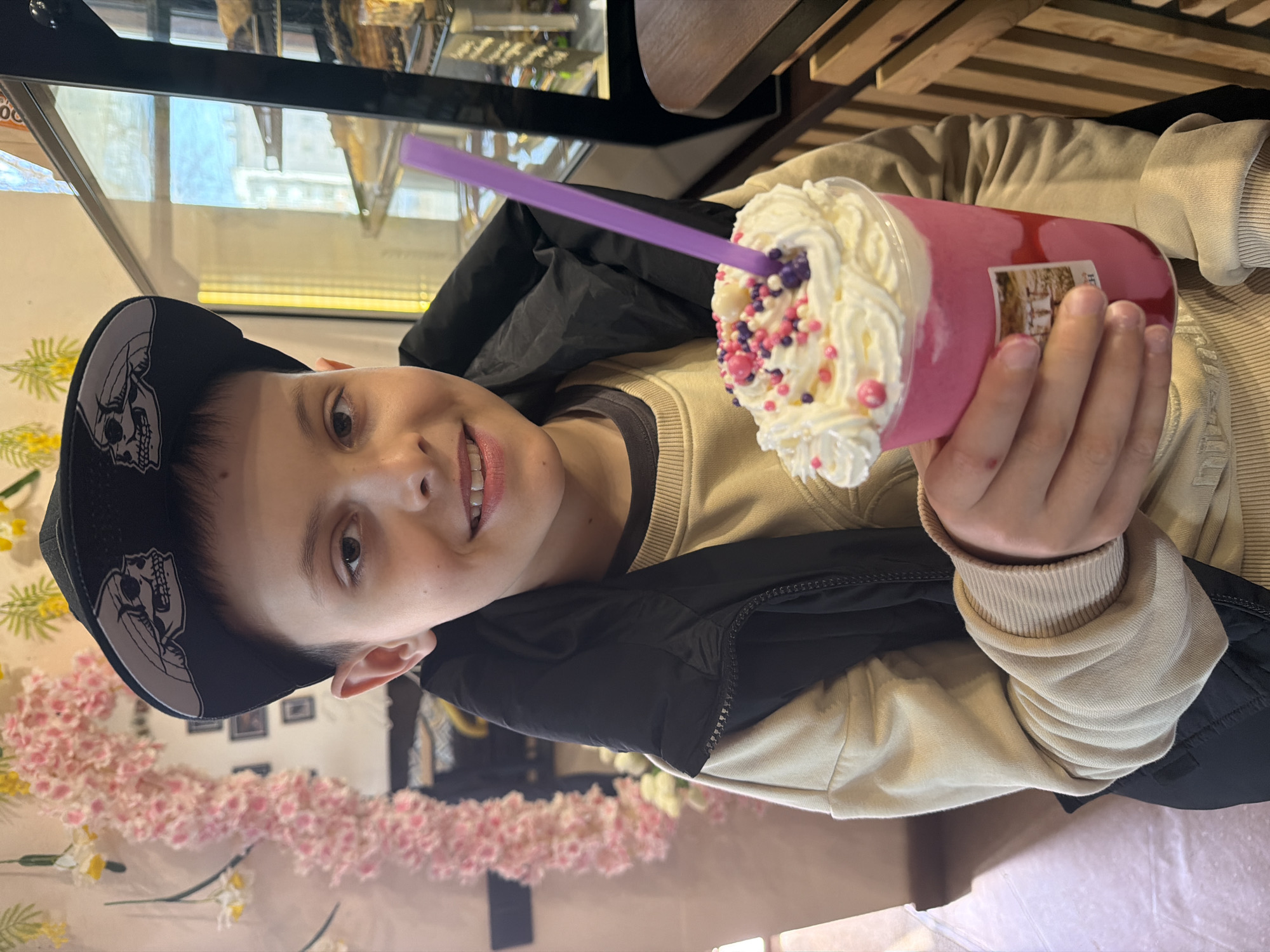 A young person, facing the camera and smiling, is holding a pink beverage with whipped cream and colorful sprinkles, along with a purple straw. The person is wearing a black cap with a skull graphic, a black and beige jacket, and a beige sweater. The background shows a display case with pastries and what appears to be a decorative floral arrangement. Based on visual cues and the mention of Votkinsk, Russia, the photo likely depicts the person inside a cafe or bakery in the city. The exact location in Votkinsk is not discernible from the image.