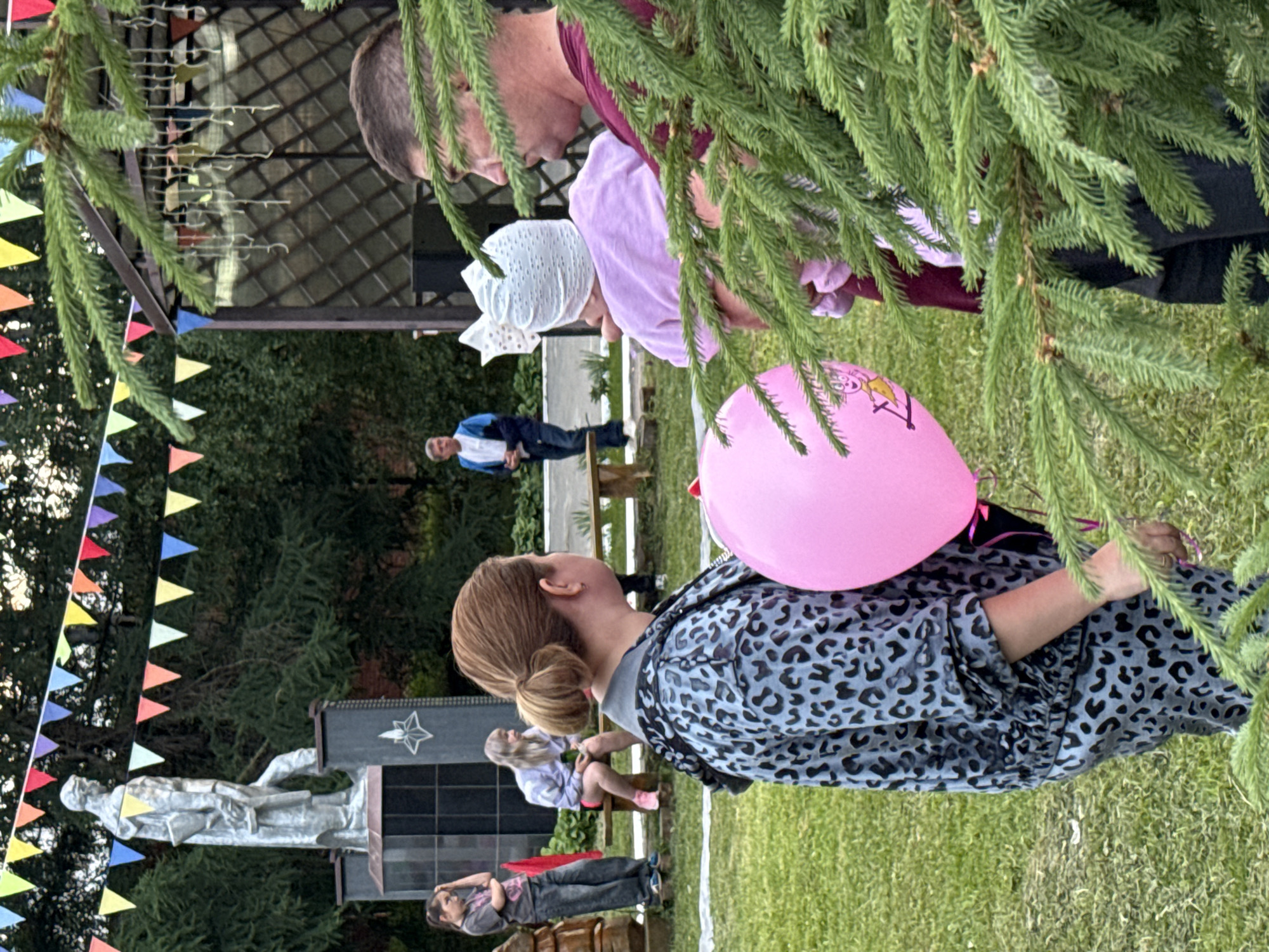 In the city of Kukui, Russia, an outdoor gathering is taking place. A woman with her hair in a bun, wearing a grey leopard-print jacket, holds a pink balloon with a cartoon character on it. She stands near a man in a dark red shirt, who is holding a baby wearing a white bow and a pink outfit. In the background, a young girl with blonde hair sits on a wooden bench, and another girl in a grey t-shirt and jeans stands nearby, resting her hand on her head. A monument with a statue is visible behind them, adorned with colorful triangular flags strung above. A gazebo with decorative elements and hanging lights is also present. The ground is covered in green grass, and a large spruce tree partially obscures the foreground.