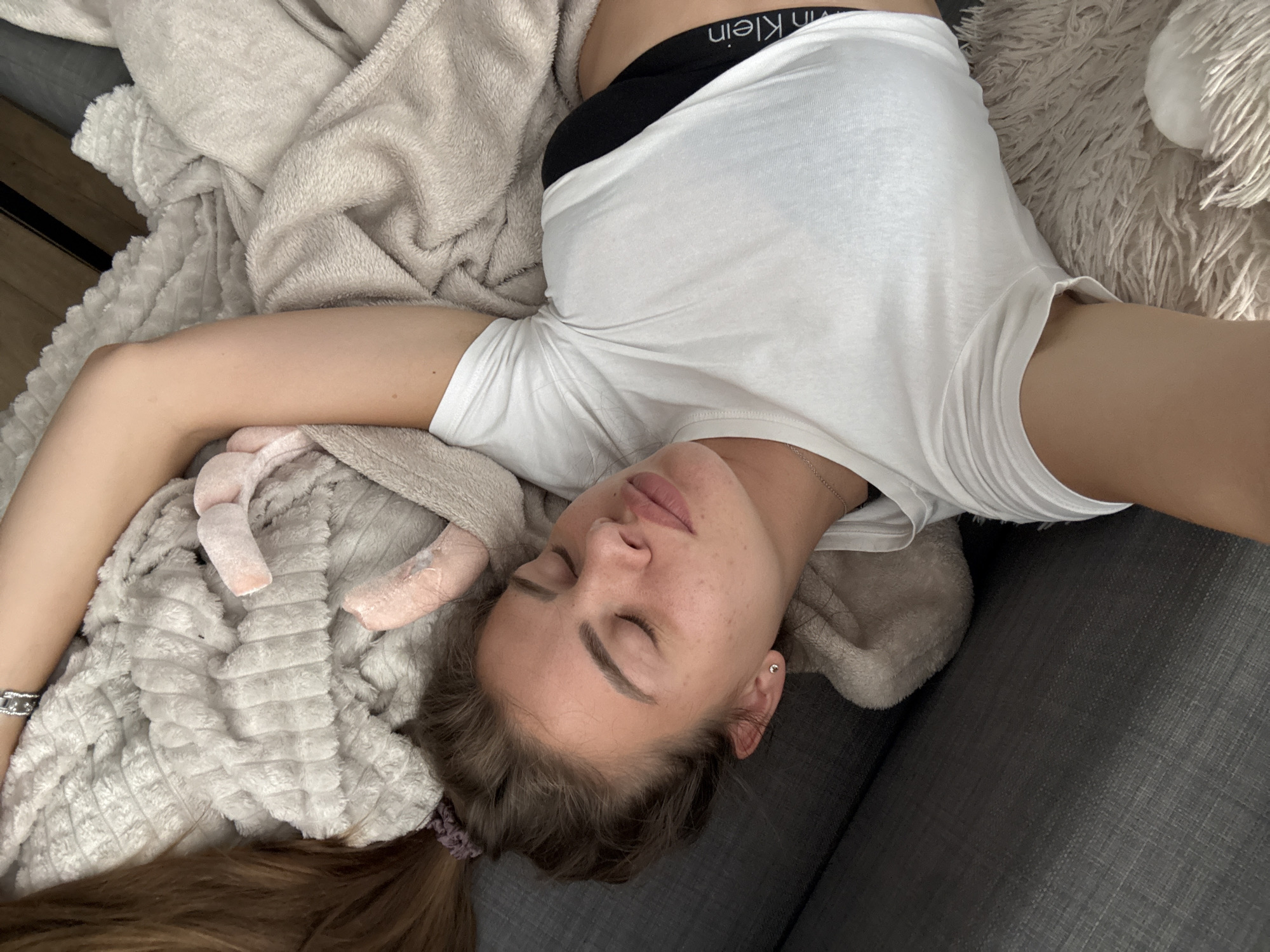 A young woman is captured resting or sleeping indoors, viewed from an overhead perspective. Her eyes are closed, and she appears calm and peaceful, lying on her back with her head slightly turned to the right. She has light brown hair, partially visible and tied back, and her face shows some freckles.
She is dressed in a loose white t-shirt, which is slightly lifted, revealing a black elastic band of an undergarment, possibly a bra or bralette, with the text "Calvin Klein" visible, oriented upside down. A thin, silver-toned necklace is around her neck. Her left arm is extended upwards, with a portion of a watch, featuring a metallic band, visible on her wrist.
The woman is partially covered and surrounded by soft, light-colored blankets or throws. One blanket has a distinct waffle-like texture, while another appears shaggy and fluffy. A small, pink, plush object is tucked close to her head, blending with the blankets. She is resting on a dark gray, textured fabric surface, likely a sofa or bed. The setting is clearly indoors, evoking a sense of relaxation and comfort.
