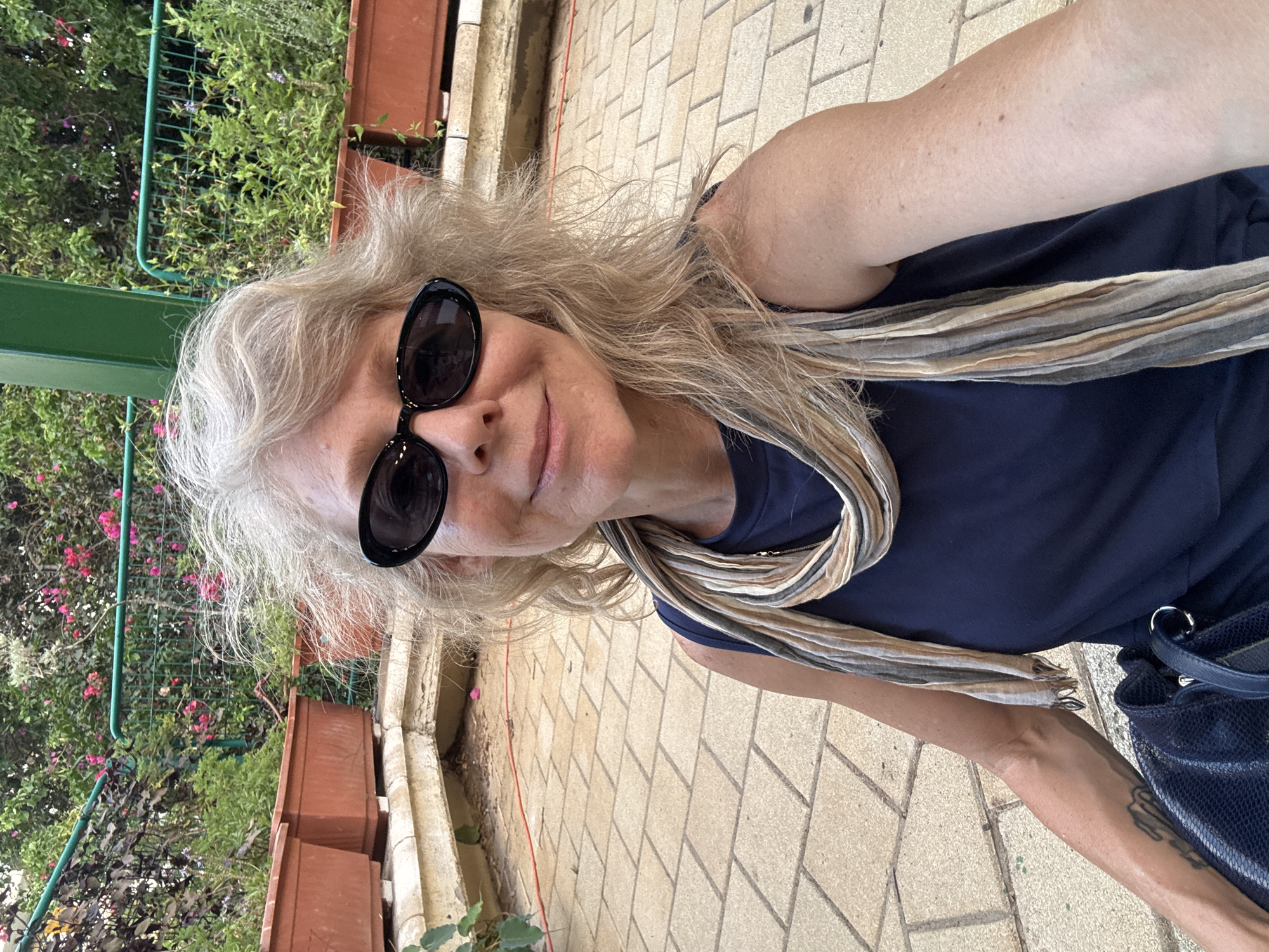 The image shows a woman taking a selfie outdoors. She has blonde, shoulder-length hair, fair skin, and is wearing dark sunglasses. She is also wearing a dark blue sleeveless top and a patterned scarf with stripes in earthy tones. A dark blue handbag is visible on her left side. The background features a paved area with tan square tiles, and behind that, several large terracotta planters containing greenery and pink flowers. A green metal fence can also be seen in the background, separating the area from more dense foliage. The lighting suggests it is daytime, possibly late afternoon or early evening due to the warm tones. The scene appears to be in a garden or park setting. There is no specific indication of the exact location within Givatayim, Israel, but the general environment suggests an outdoor public space.