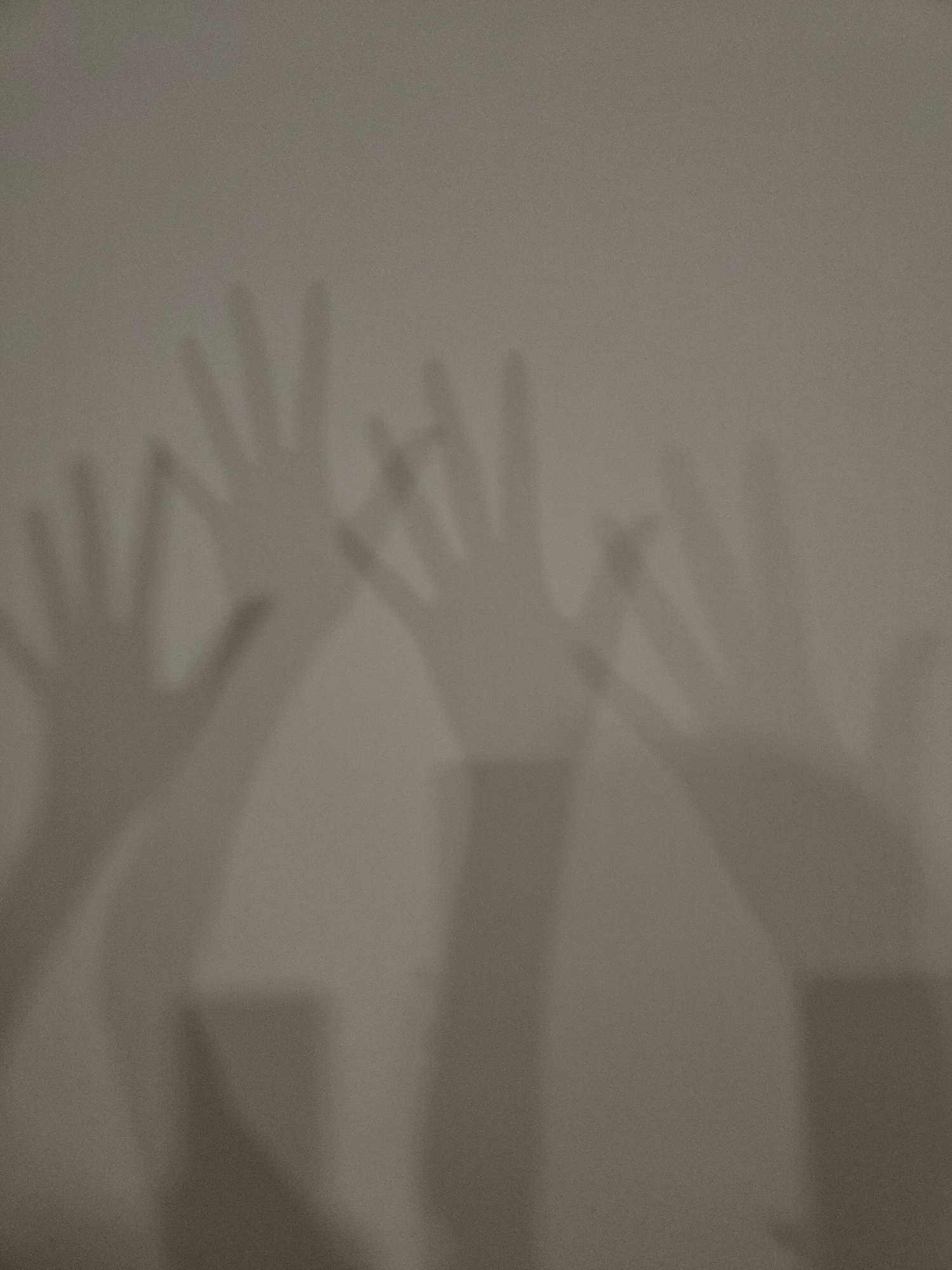 The image captures multiple shadows of human hands cast upon a plain, light-colored, textured surface, likely a wall. There are at least three distinct hand shadows visible, with their fingers splayed and extended upwards. One shadow on the far left shows a hand and part of an arm extending downwards. In the central area, two hand shadows appear to be overlapping or close together, and another hand shadow is present on the right, also showing an arm. The light source creating these shadows is soft and diffused, resulting in gentle, rather than sharp, shadow edges. The background surface is a consistent shade of light gray or beige and has a subtle, fine-grained texture throughout. The scene appears to be an indoor setting, possibly depicting a playful or artistic moment involving individuals casting hand shadows. There are no other discernible objects or visible text in the image.