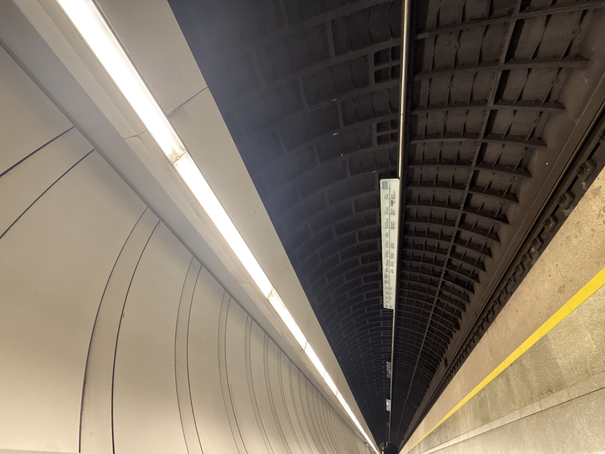 This image shows a view of the Park Pobedy metro station in Moscow, Russia. The architecture features curved walls on the left, illuminated by a line of lights. The ceiling on the right side has a dark, patterned design. A yellow safety line runs along the edge of the platform. A sign hangs from the ceiling. There are no people visible in the scene.