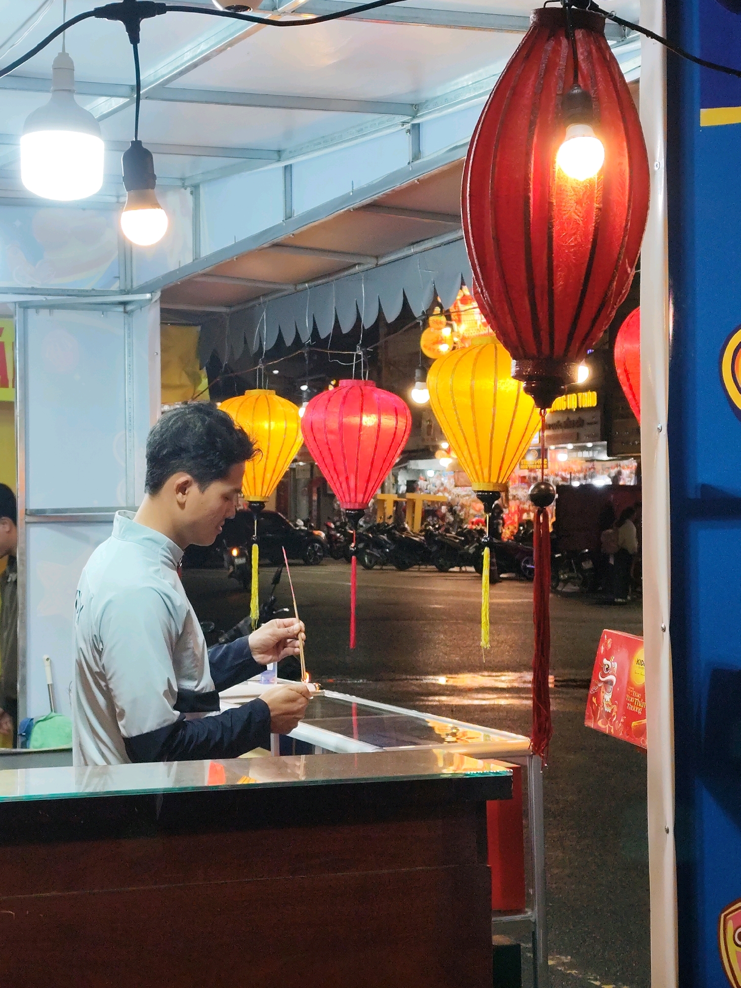 The image depicts a man standing at a stall in Da Lat, Vietnam, at night. He is holding two incense sticks, one of which is lit, and appears to be preparing them. The stall is decorated with various colorful lanterns, including red and yellow ones, which are illuminated by internal bulbs. In the background, a street scene is visible with motorbikes parked and people moving about. The overall ambiance suggests a lively night market or street-side establishment. The stall is framed by metal poles and has a wooden counter. The man is wearing a light-colored jacket with dark sleeves.