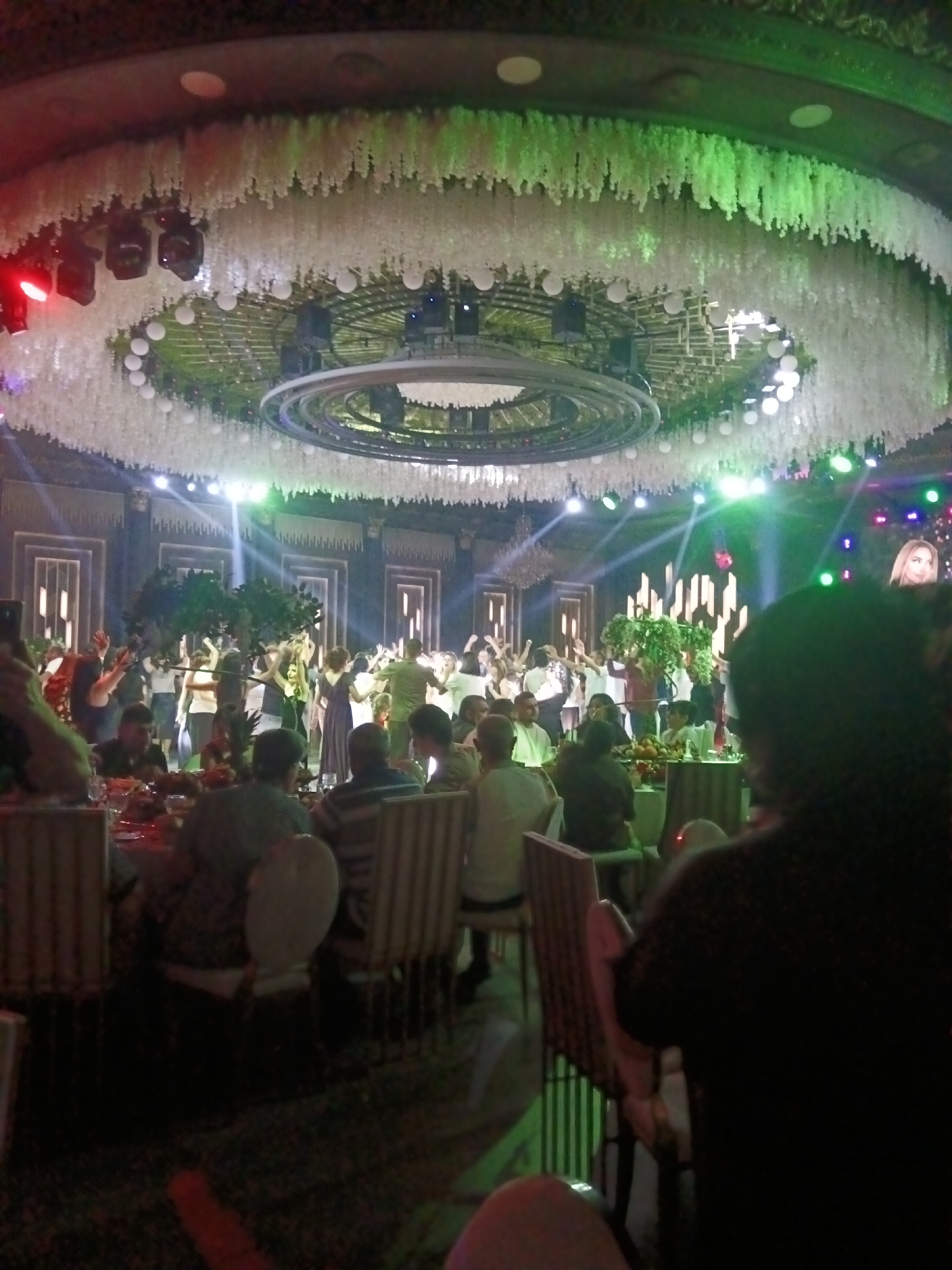 The image depicts a lively indoor event, possibly a wedding reception or a large party, at the Etchmiadzin Mother See complex in Vagharshapat, Armenia. A large gathering of people are visible, many dancing or mingling in the central area of the venue. The ceiling is elaborately decorated with cascading white floral arrangements and suspended lighting fixtures, some of which cast green and red light onto the scene. In the background, a stage area is set up with vertical decorative elements and spotlights. A large screen displays a portrait of a woman. Tables with food and drinks are set up in the foreground, where some guests are seated and others are partially visible. The overall atmosphere appears festive and energetic.