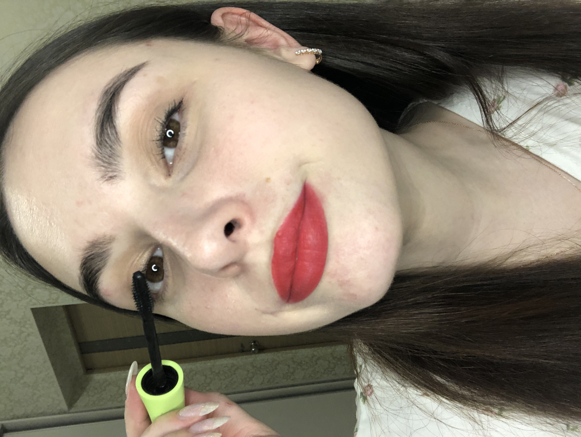 A fair-skinned woman with brown hair and light brown eyes is applying mascara to her lower eyelashes. She is wearing red lipstick. Her nails are long and appear to have glitter polish. She has a small hoop earring with crystals in her left ear. The background appears to be an interior space, possibly a bathroom or bedroom, with light-colored walls. The city location in Noviy, Russia is extraneous.