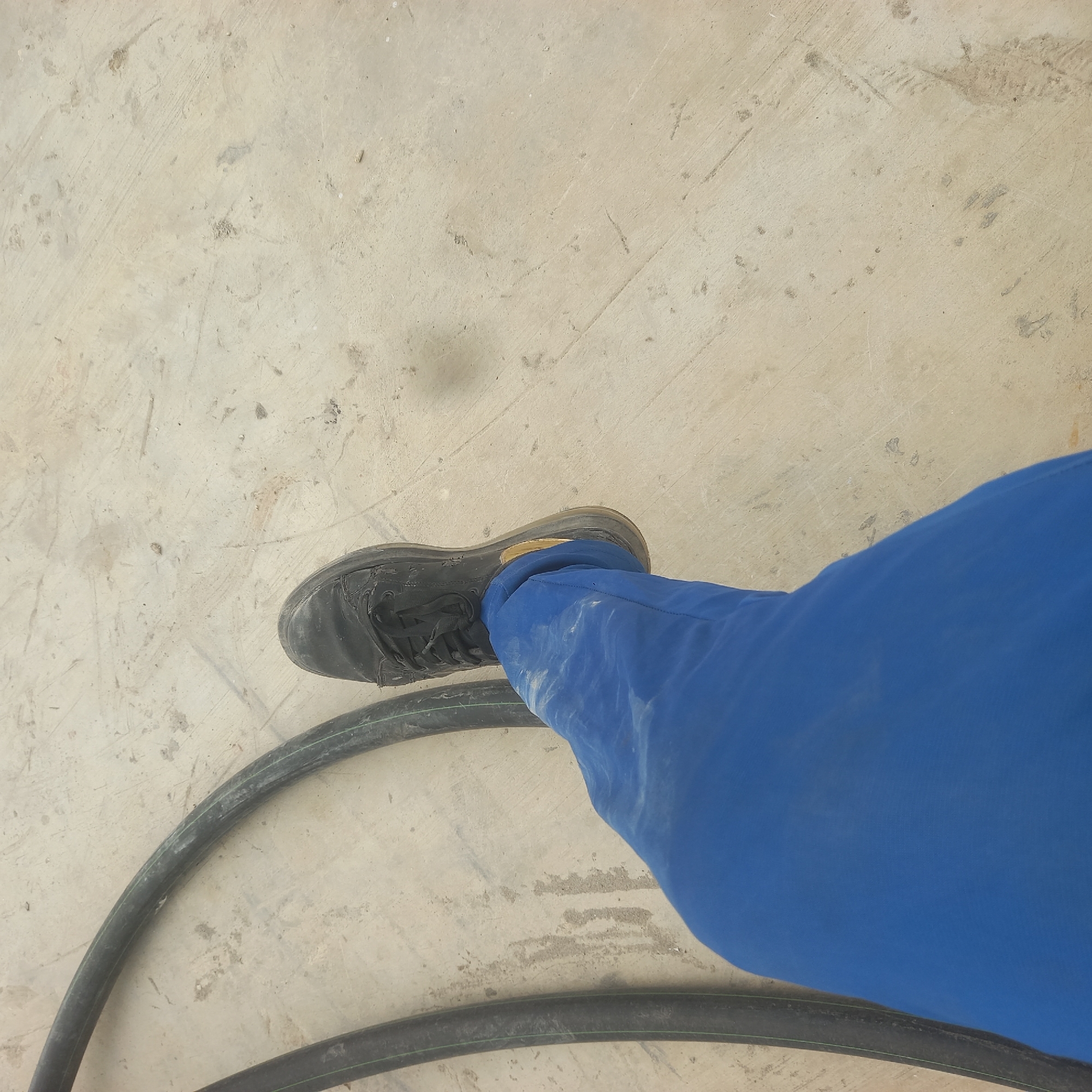 A person's left leg, clad in bright blue trousers covered in white dust, is visible from the knee down. The leg is positioned to the left of the frame, with the foot shod in a black, scuffed sneaker. The sneaker has laces and appears to be worn. Beside the leg, a thick, black hose with a subtle green stripe runs in a curved path across the light-colored, dusty ground. The ground itself is concrete or similar material, marked with faint lines and scattered debris, consistent with an outdoor or construction environment. The scene is devoid of any identifiable city features or landmarks that would specifically place it in Bububu, Tanzania.