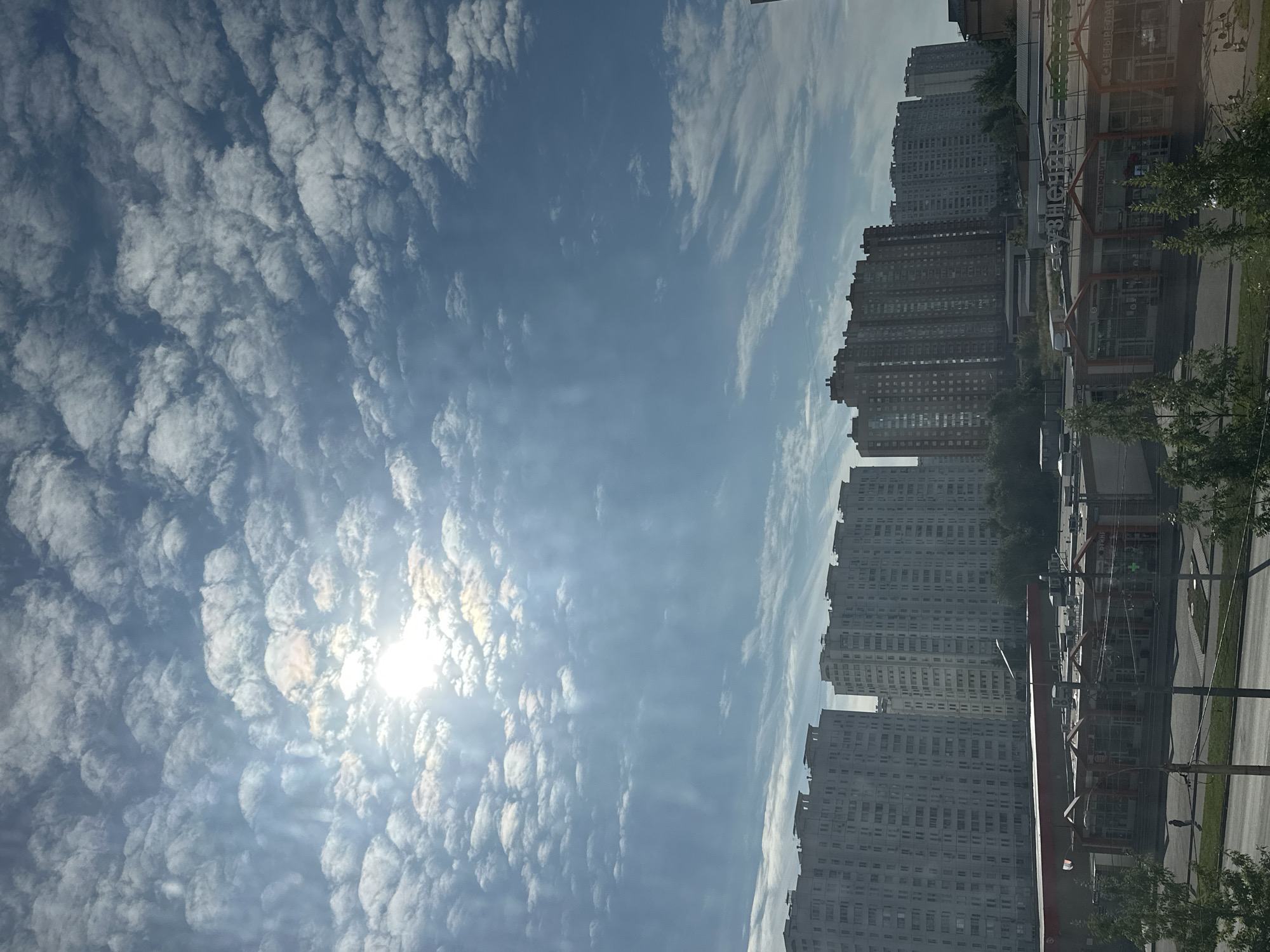 The image depicts a bright, sunny day in Podolsk, Russia, with a clear blue sky filled with scattered cumulus clouds. The sun is high in the sky, casting a bright light and creating sunbeams that stream through the clouds. In the foreground, a row of multi-story apartment buildings is visible, with a shopping center situated in front of them. The shopping center features large windows and an awning, and its facade is adorned with various signs, including one that reads "УЗНЕЧКА" (Uznechka) and another that appears to be a part of a supermarket's name. Small trees are planted in front of the shopping center, and a road runs parallel to it. The overall scene suggests a typical urban residential and commercial area on a pleasant day.
