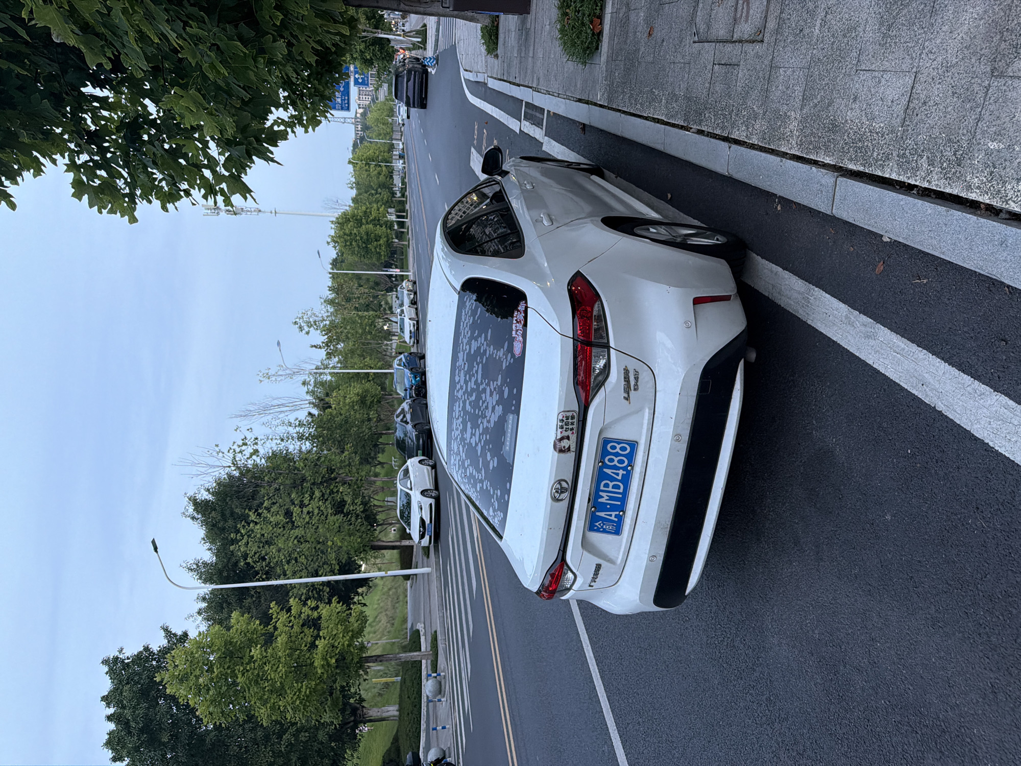 In the city of Jiangjin, China, a white sedan, identified by the license plate A-MB488, is parked on the right side of a paved road. The car is viewed from a rear-quarter angle, with its left side closest to the camera. The vehicle's rear window is partially obscured by a pattern of white dots, possibly stickers or fading. A small sticker with a cartoon image of a woman and text is visible on the lower left side of the rear bumper. The road features double yellow lines and white lane markings. Across the street, a pedestrian crossing with white stripes is marked, and several other vehicles are driving or parked. The environment includes a sidewalk with granite paving stones and a curb separating the road from a grassy area with trees. Streetlights and communication towers are visible in the background, against a cloudy sky. The scene appears to be a street in a developed urban or suburban area.